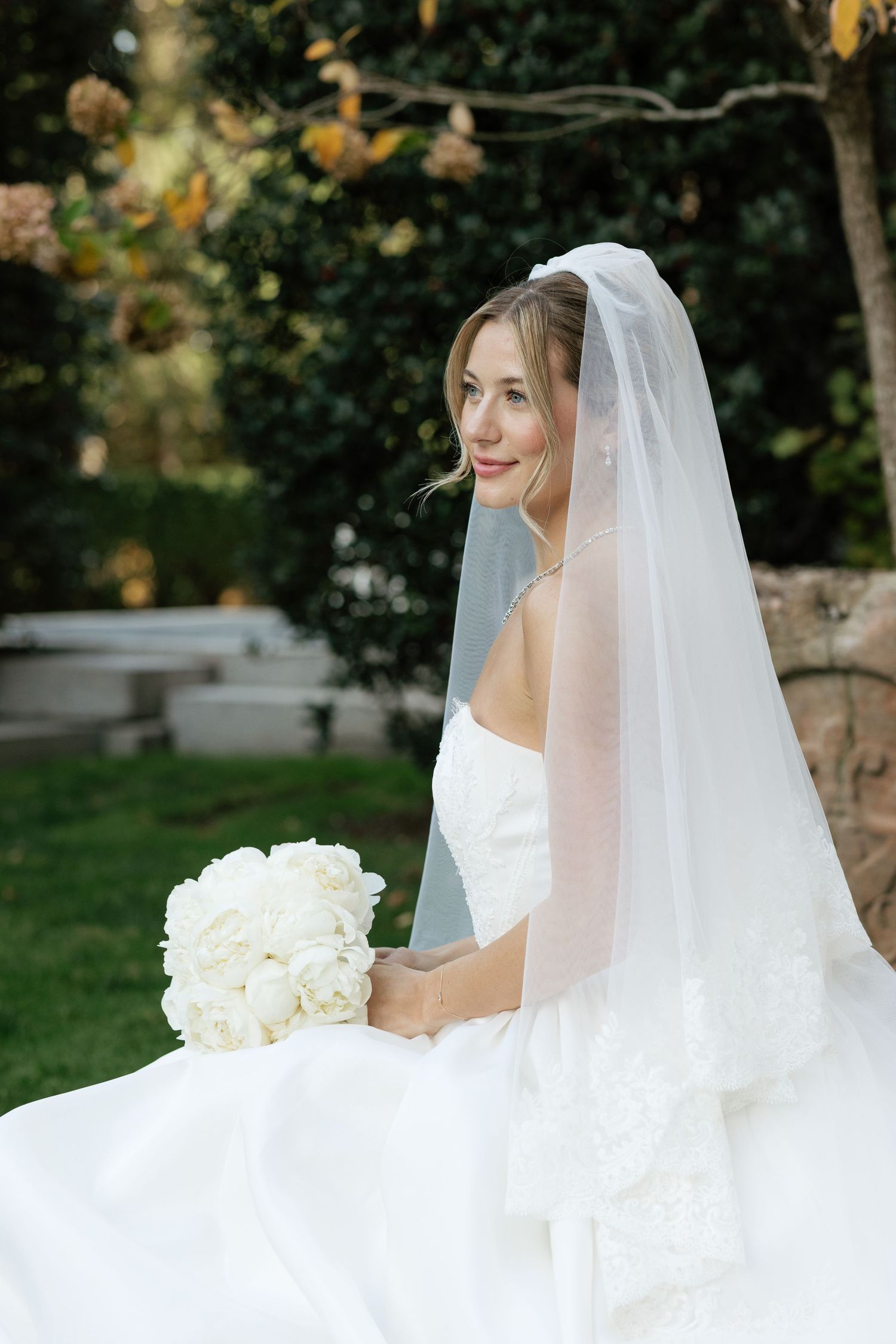 Bride in white wedding gown and veil poses with white roses in a garden setting.
