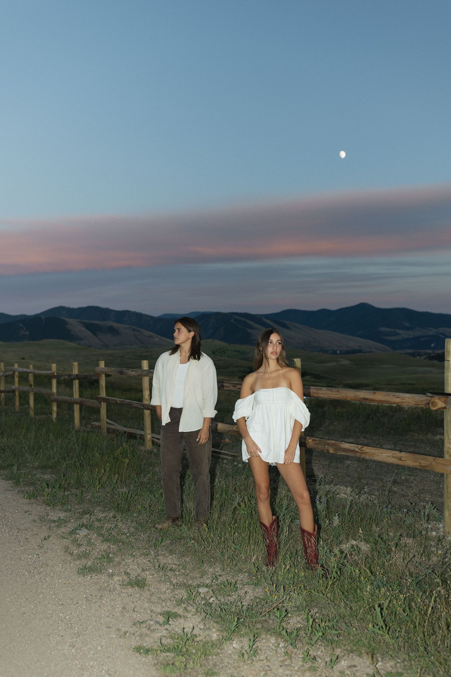 Couple in casual attire stands by wooden fence at sunset overlooking mountainous landscape.