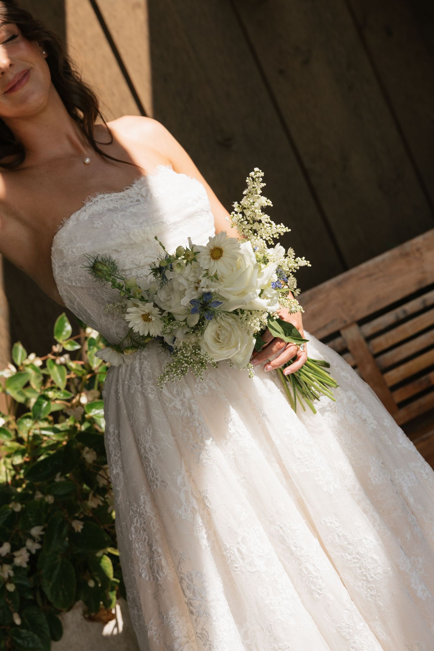 Bride in strapless lace wedding gown holding a white and blue wildflower bouquet with roses and daisies outdoors.