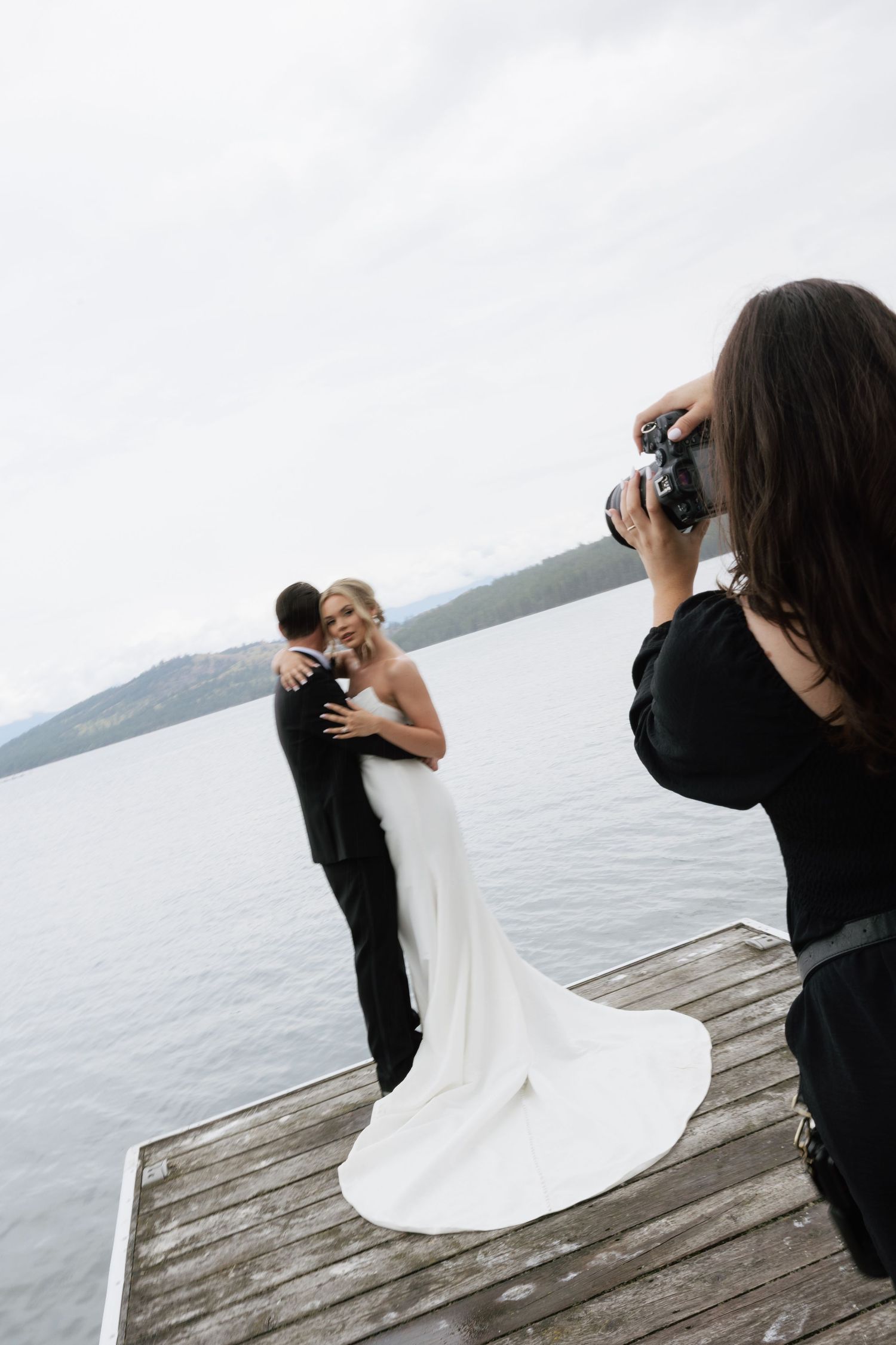 Wedding photographer capturing bride and groom embracing on a wooden dock overlooking a serene lake and mountains.