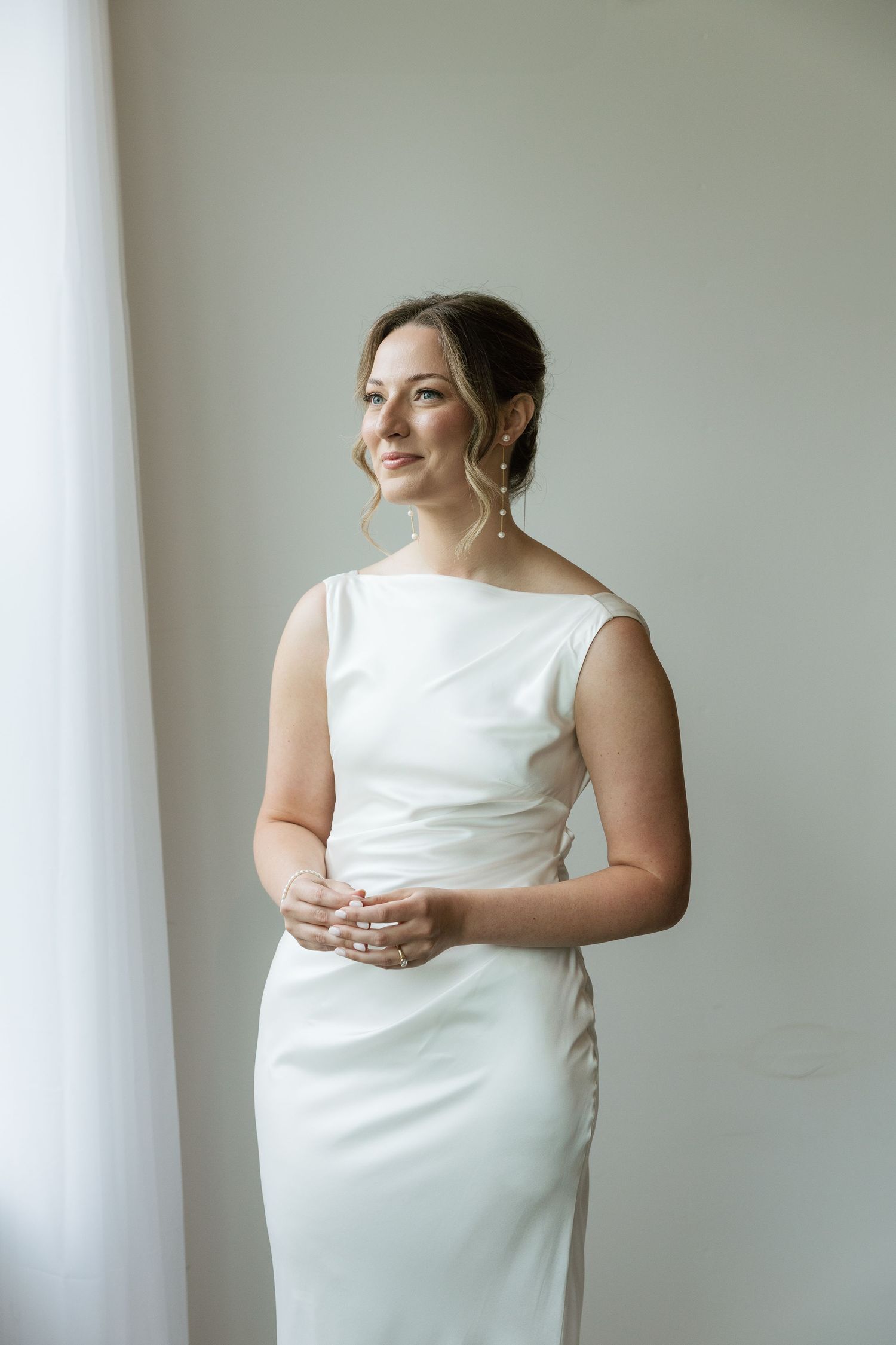 Elegant bride in a sleek white sleeveless wedding gown standing by a window, smiling softly with hands clasped.