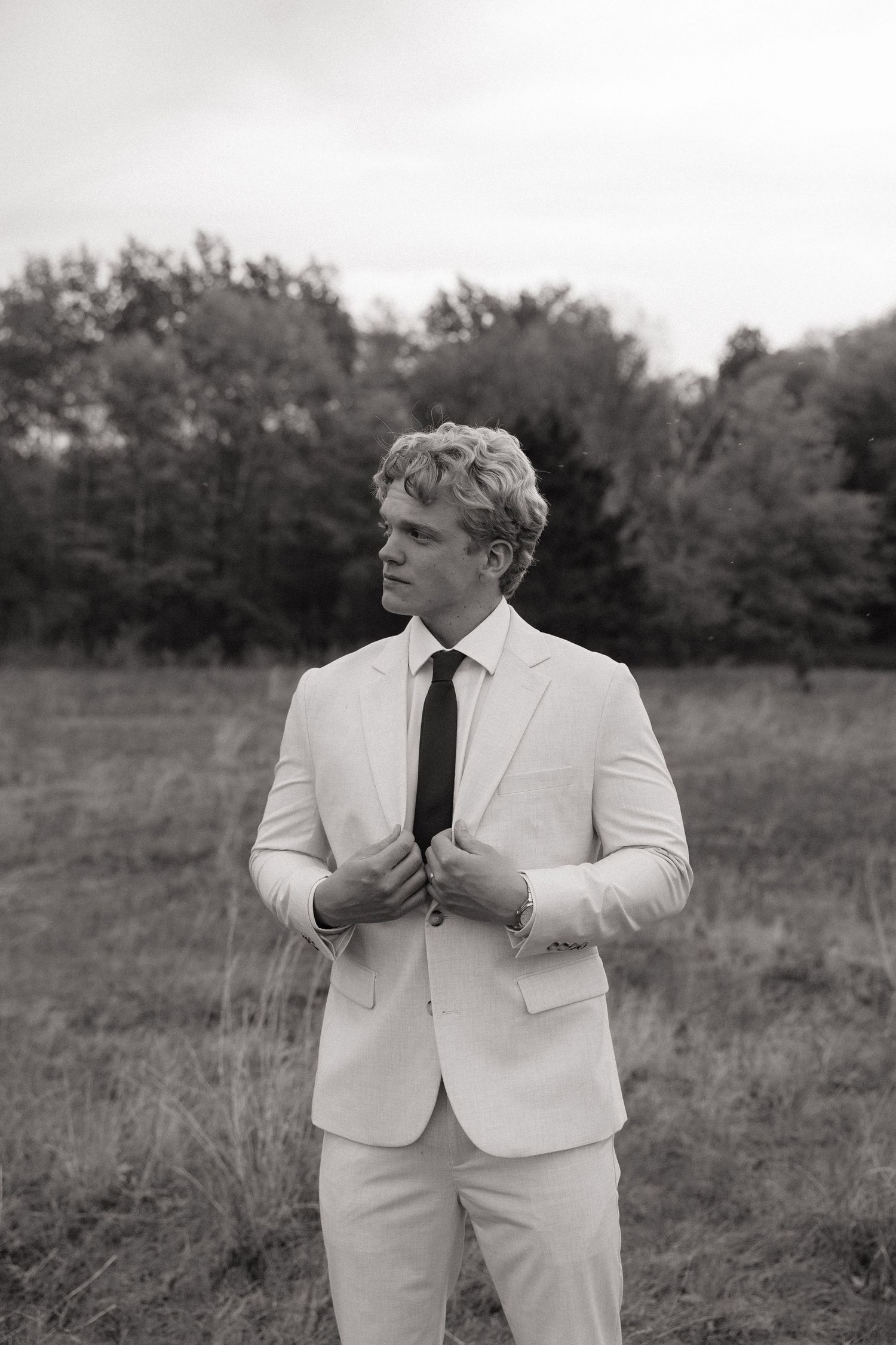 Black and white photo of a young man in a light suit and dark tie standing in a field with trees in the background.