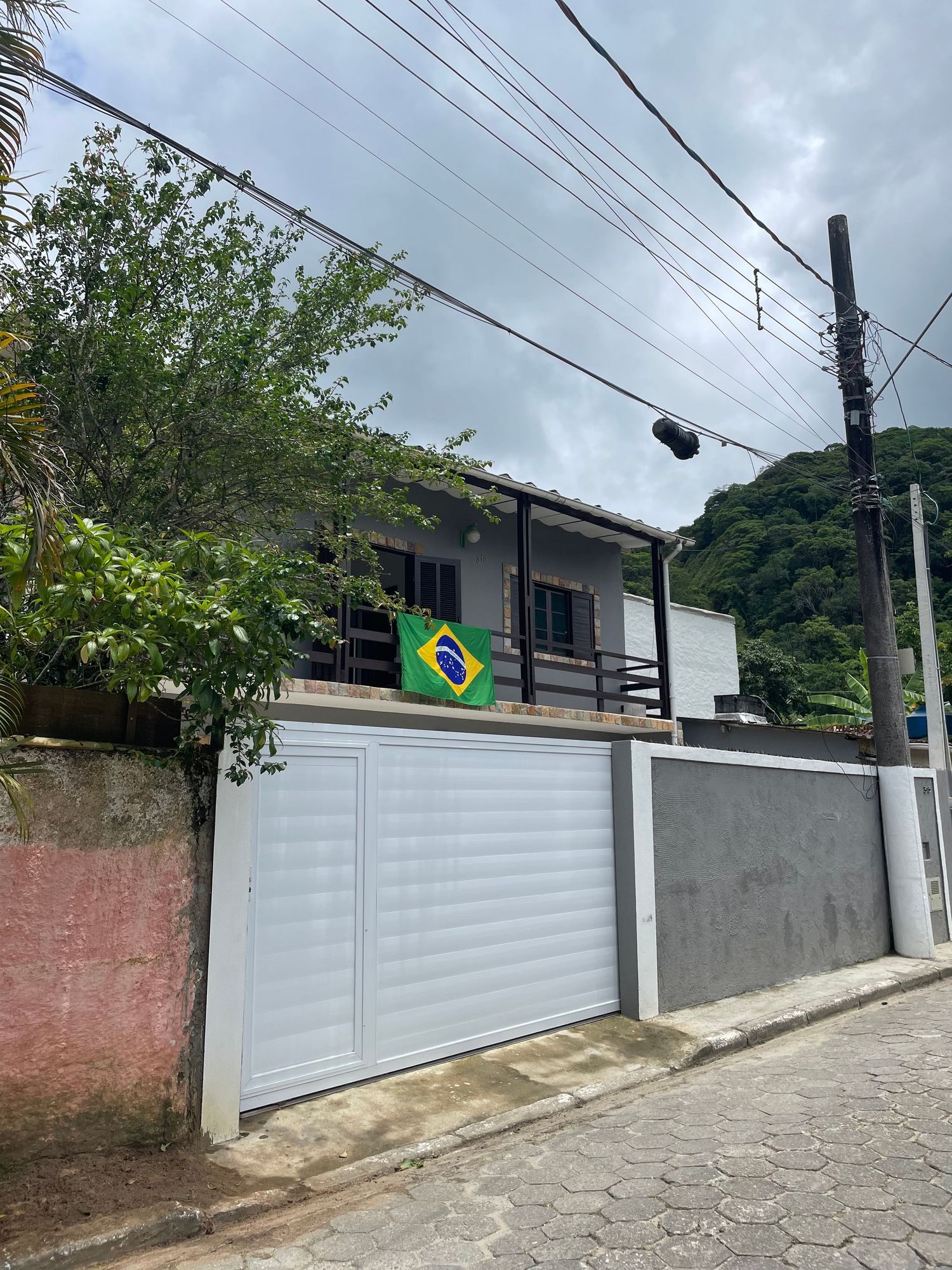 Two-story house with white garage door and Brazilian flag displayed on balcony, surrounded by power lines and trees.