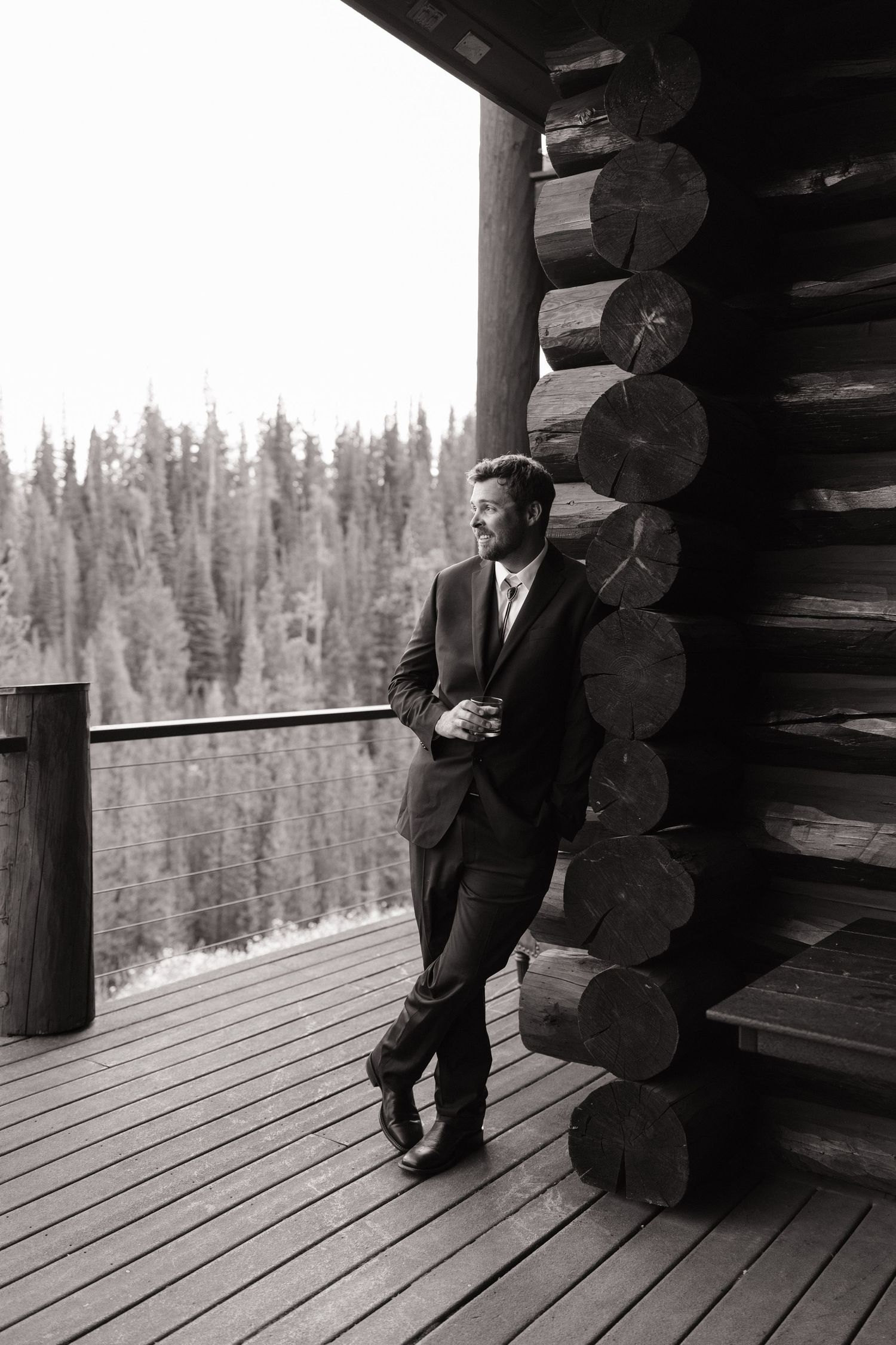 Black and white image of a rustic log cabin porch overlooking a forest landscape.