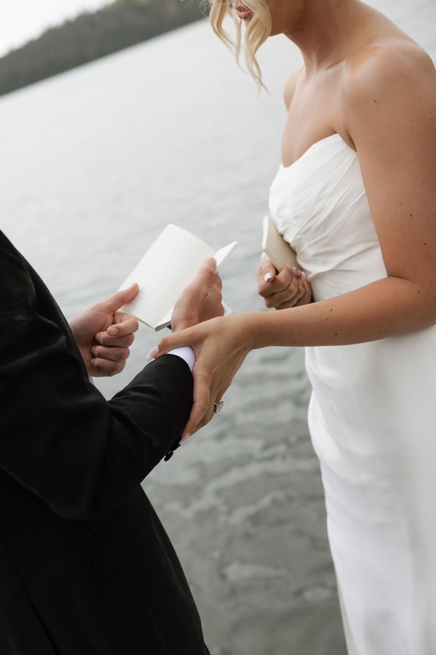 Groom reading wedding vows while holding bride's hand in white strapless gown by a lakeside ceremony.