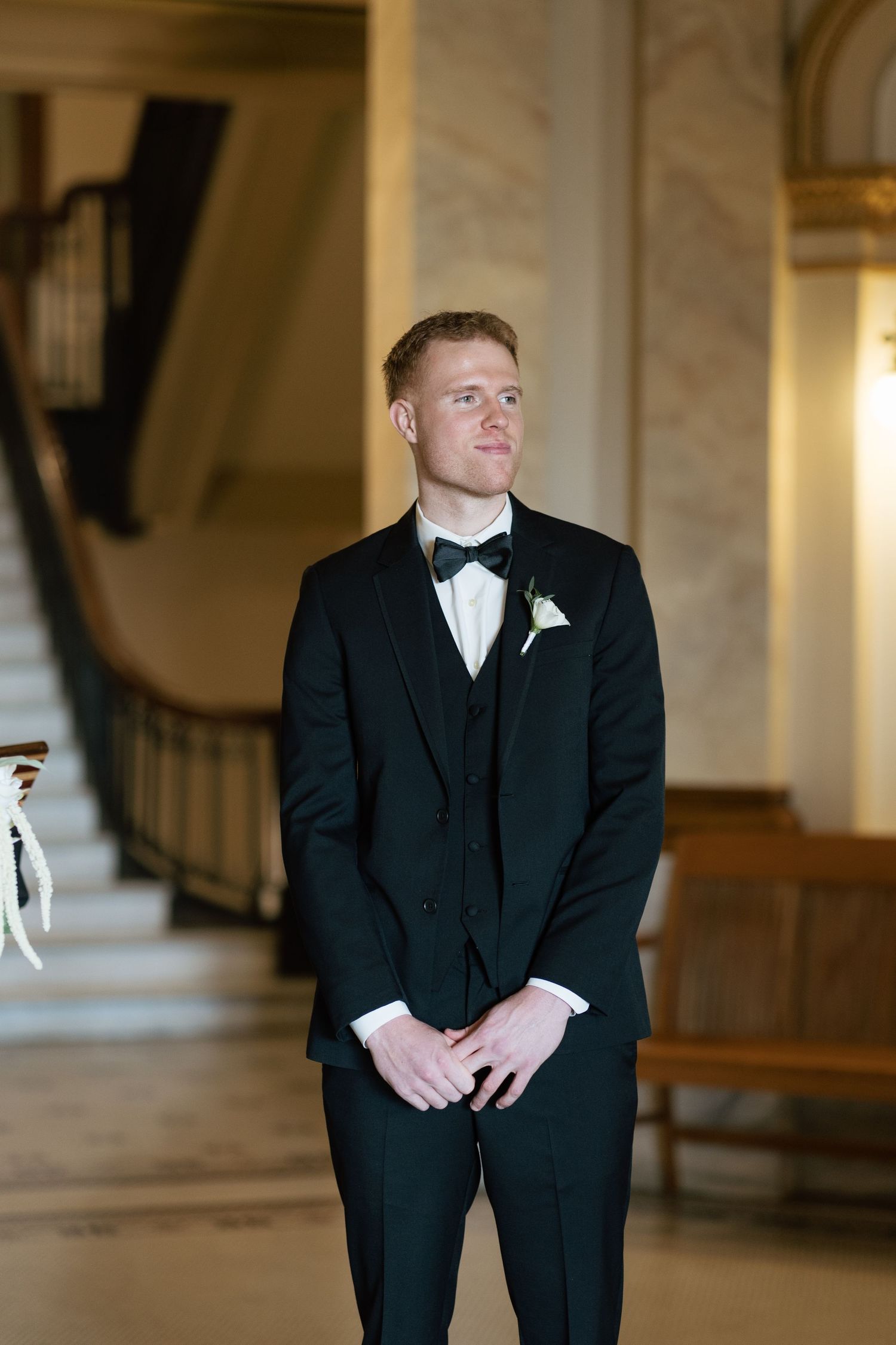 Groom in classic black tuxedo poses in elegant marble hallway.