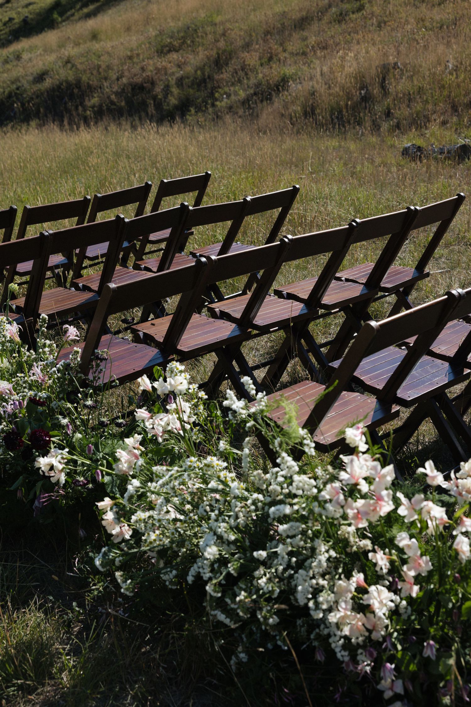 Wooden ceremony chairs decorated with lush white floral arrangements on a hillside.