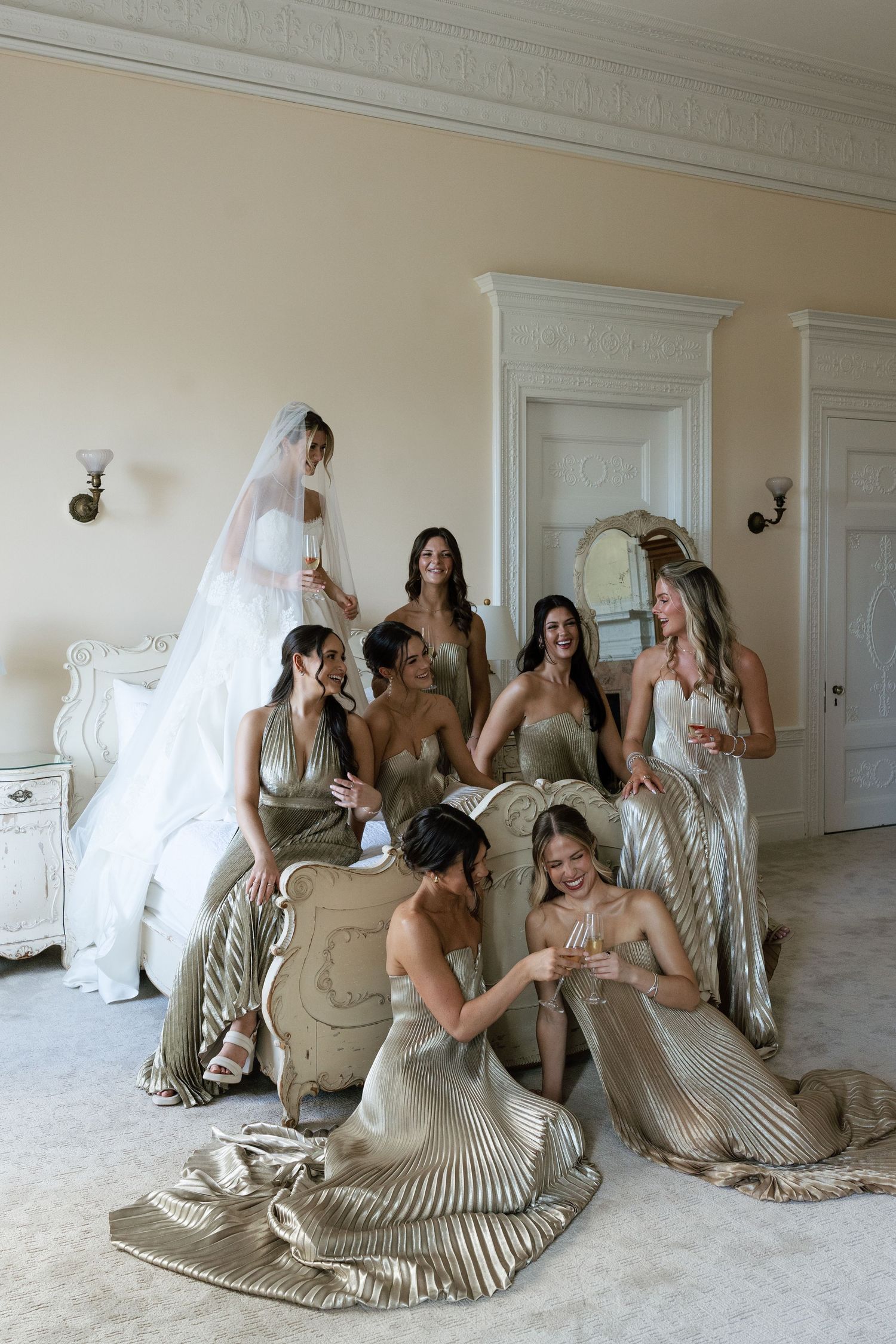 Wedding party in champagne-colored dresses sitting together in an elegant white room.