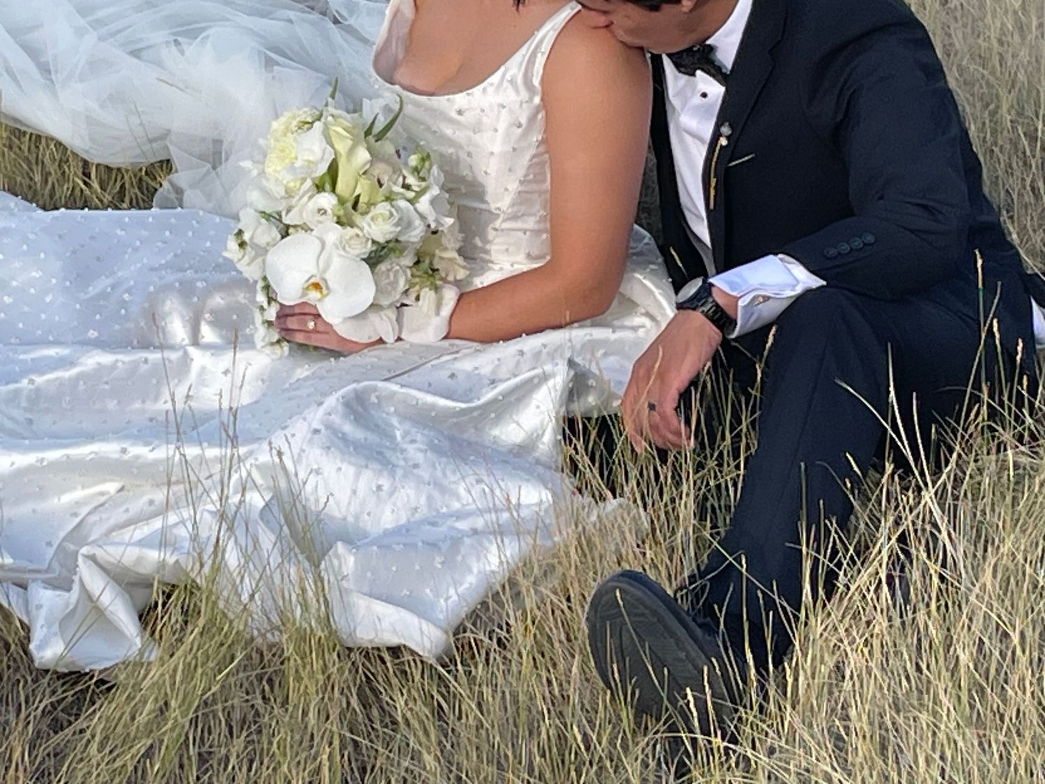 Bride and groom sitting in golden grass field, bride in white gown holding bouquet, groom in black tuxedo.