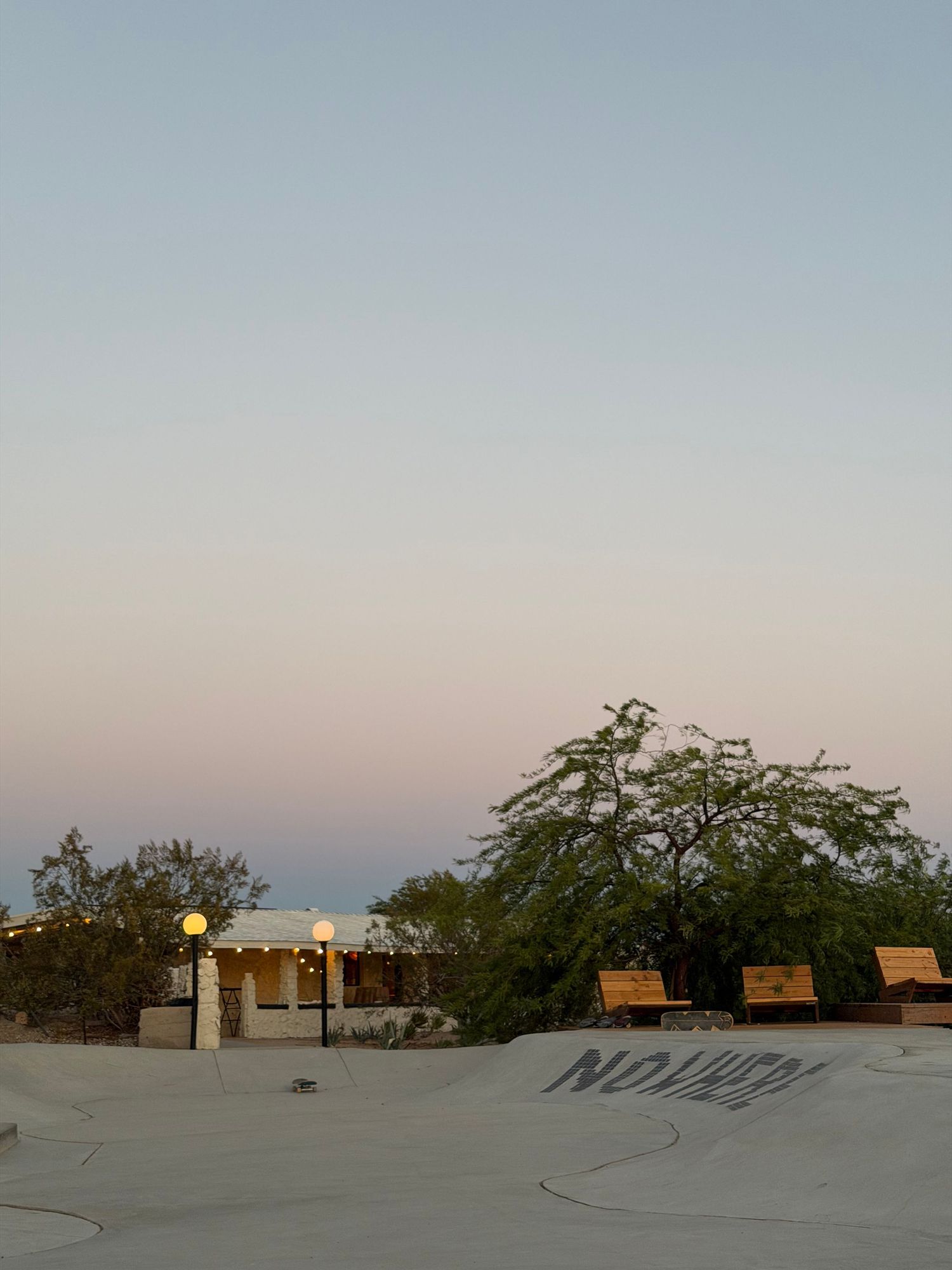Skate park ramp at dusk with a small building, benches, and green trees under a pastel pink and blue sky.