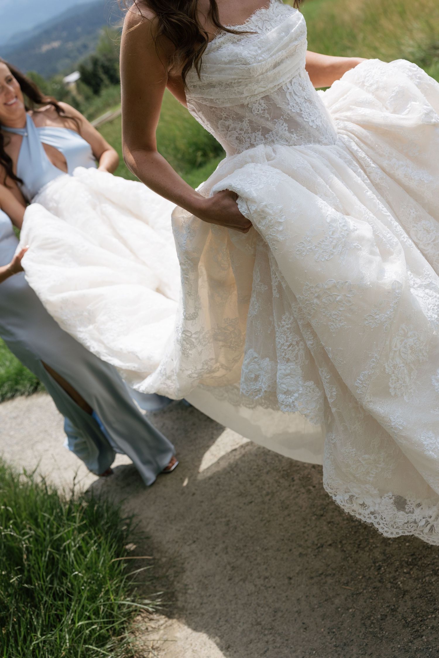 Bride in lace ballgown wedding dress being helped by bridesmaid in silver dress outdoors on a sunny day.