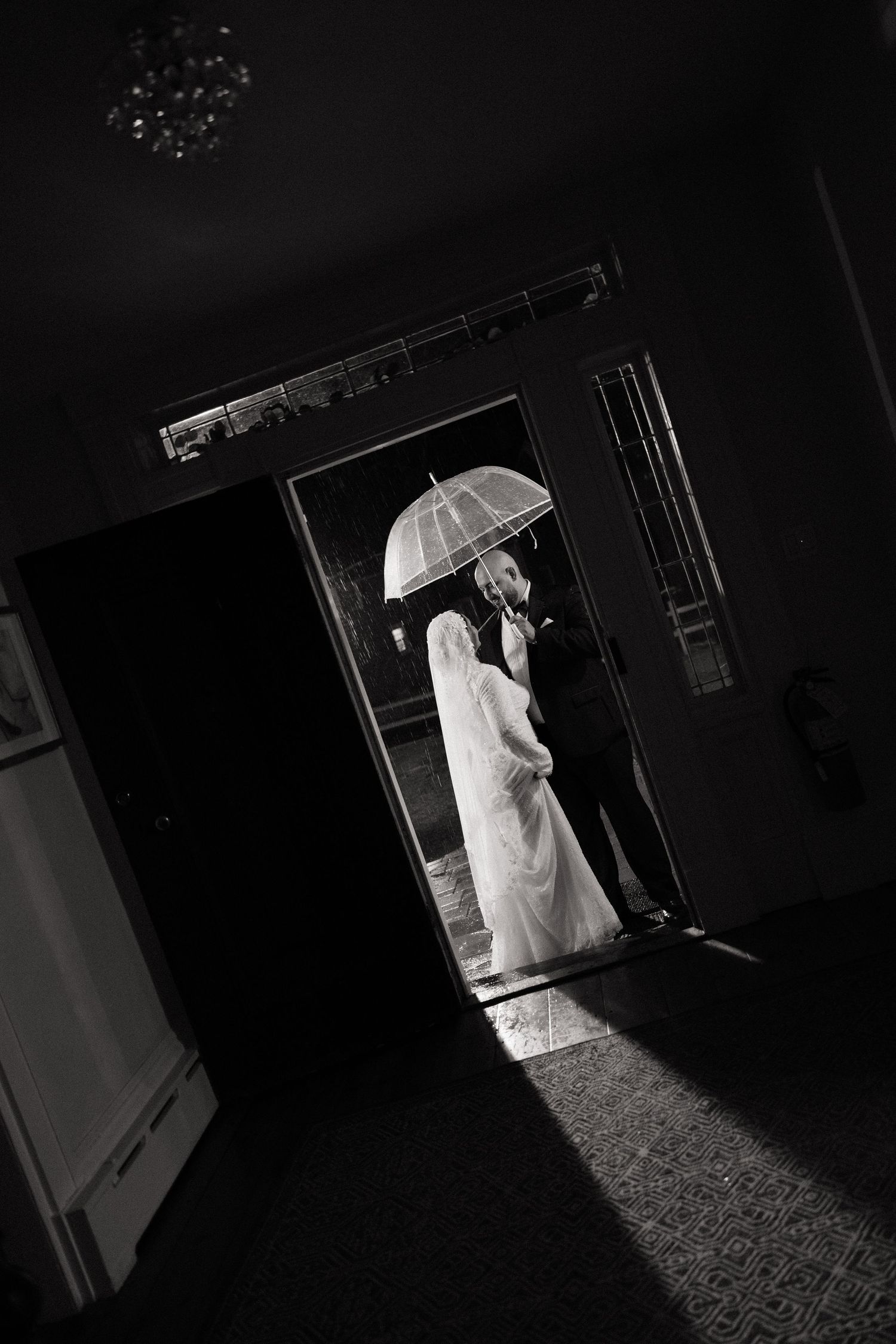 Black and white wedding photo of bride and groom sharing umbrella in doorway, romantic rainy day portrait.