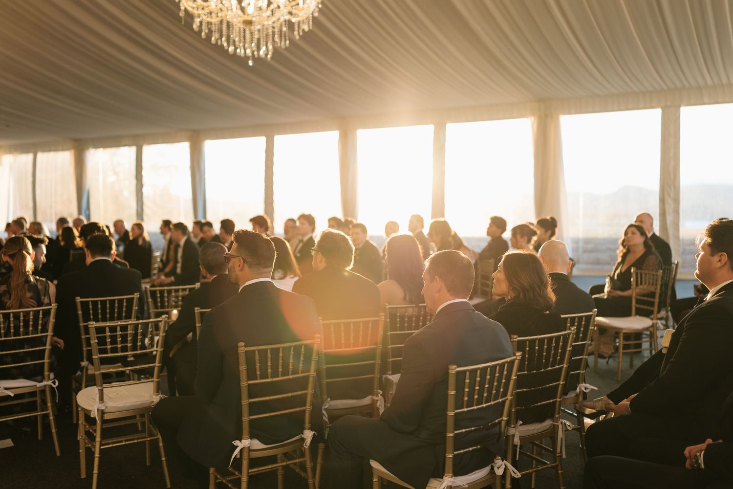 Wedding ceremony setup with chairs and guests backlit by golden sunset through tent.