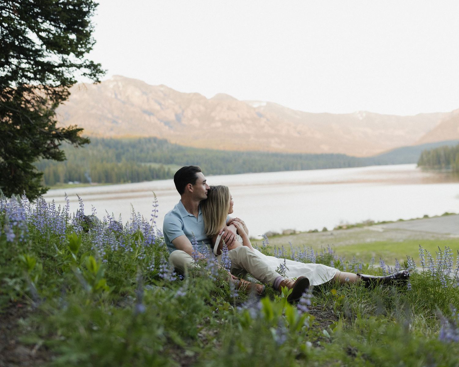 A couple shares an intimate moment on a hillside overlooking a serene lake with mountains in the background.