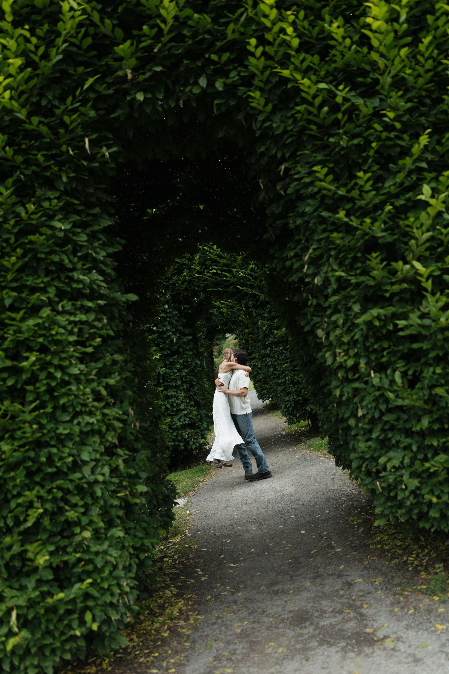 A couple embracing under a natural archway formed by green hedges on a garden path.
