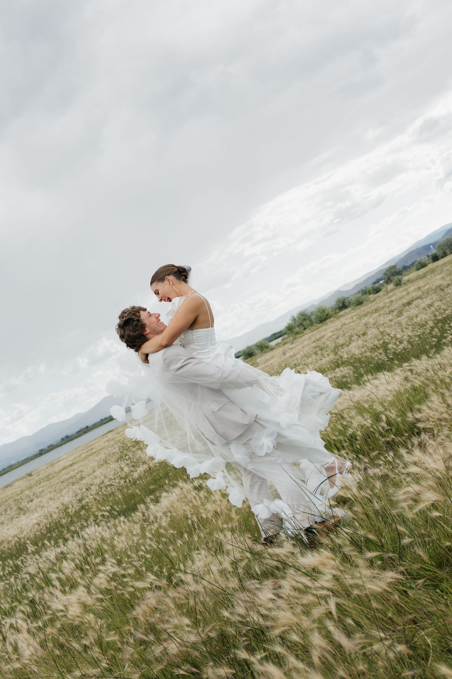 A bride in a flowing white dress spins gracefully in a windswept meadow against a cloudy sky.