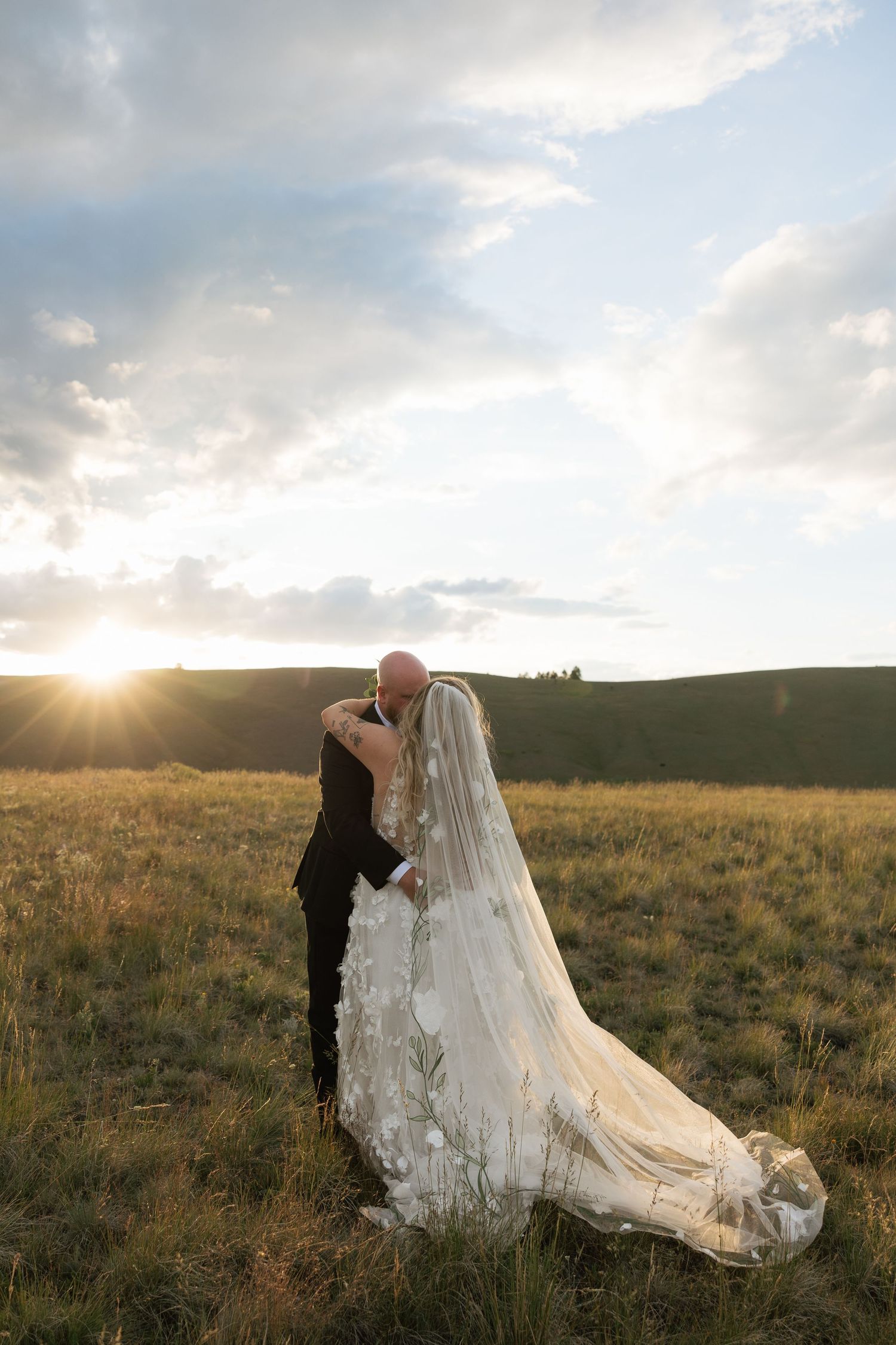 Bride and groom kissing at sunset in a golden field, her floral cathedral veil trailing gracefully on the grass.