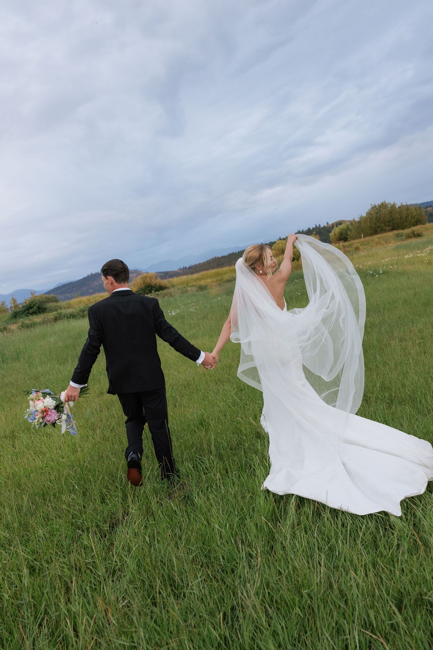 Bride and groom walk hand-in-hand through green meadow, her veil flowing in wind, mountains in background.