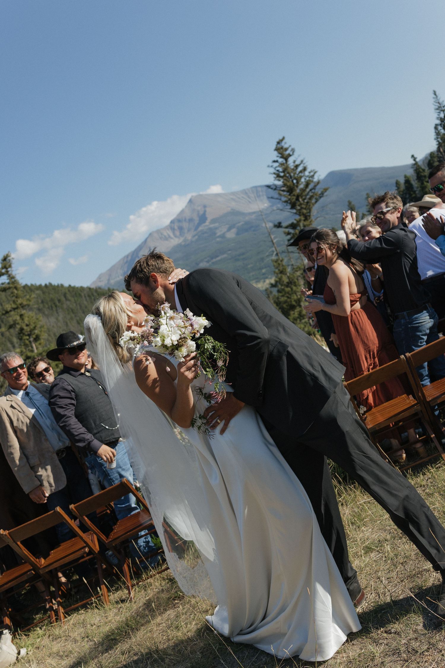 Outdoor mountain wedding ceremony with guests seated on wooden chairs against scenic backdrop.