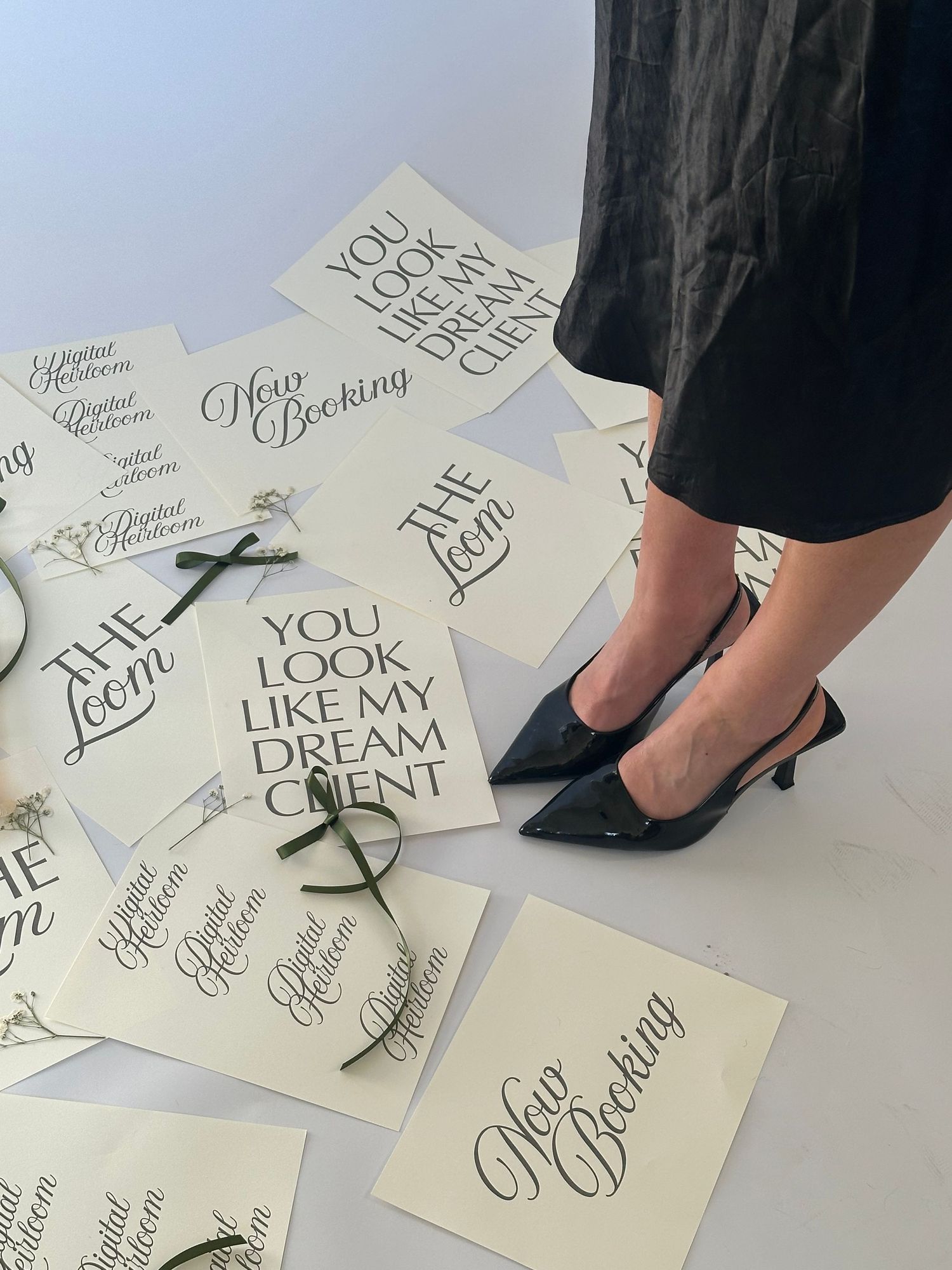 Elegant calligraphy cards with phrases like 'You Look Like My Dream Client' and 'Now Booking' scattered on floor near black heels.