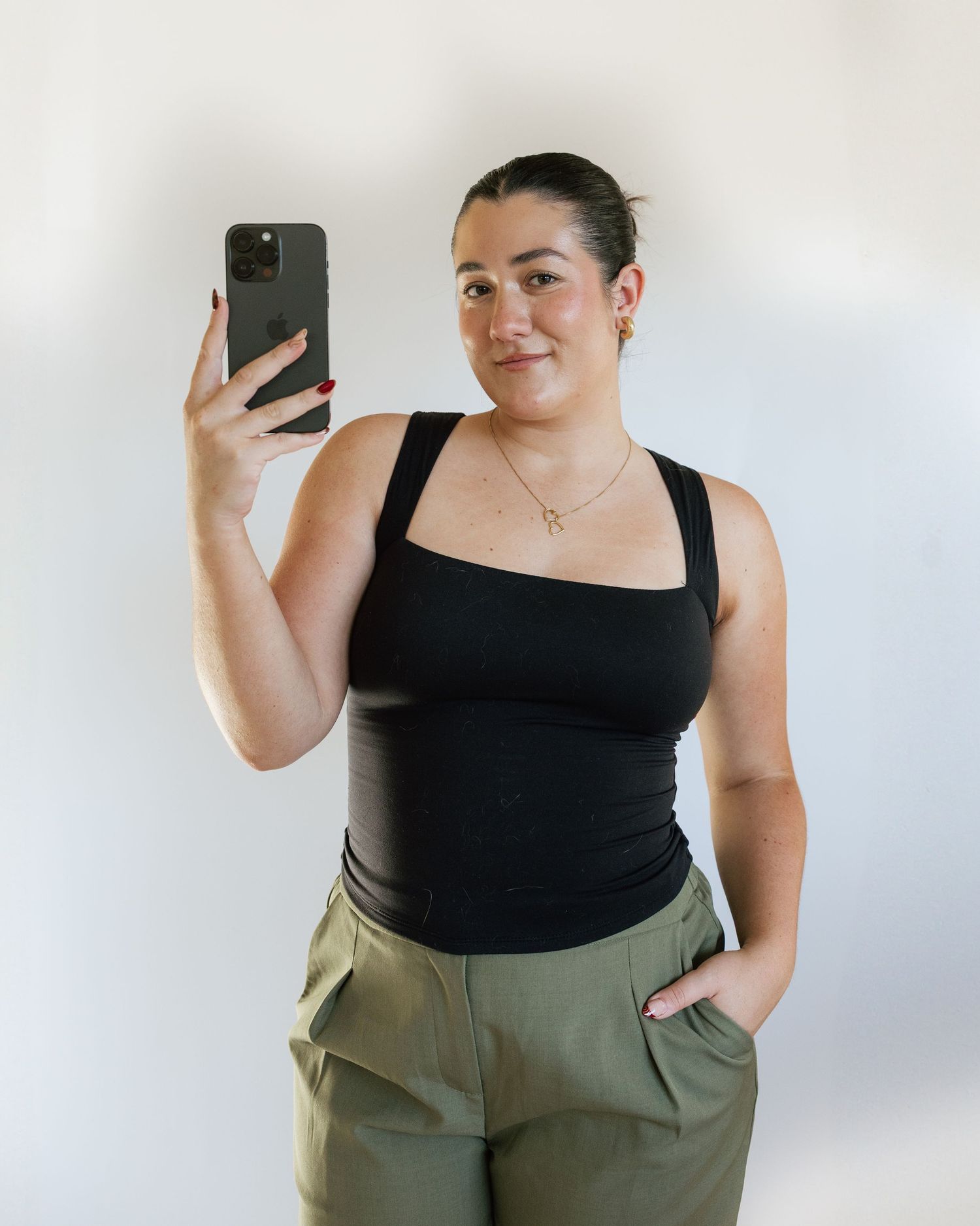 Woman in black tank top and olive green pants taking a mirror selfie with a smartphone.