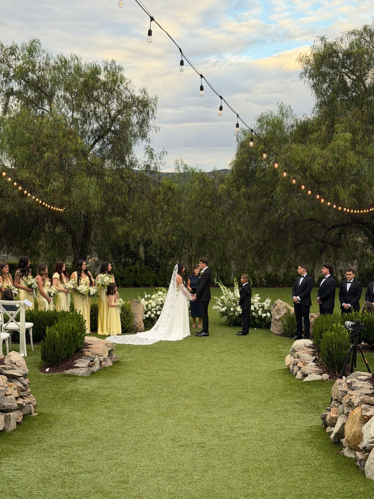Outdoor wedding ceremony on lush green lawn with string lights, yellow bridesmaid dresses, and floral arrangements at dusk.