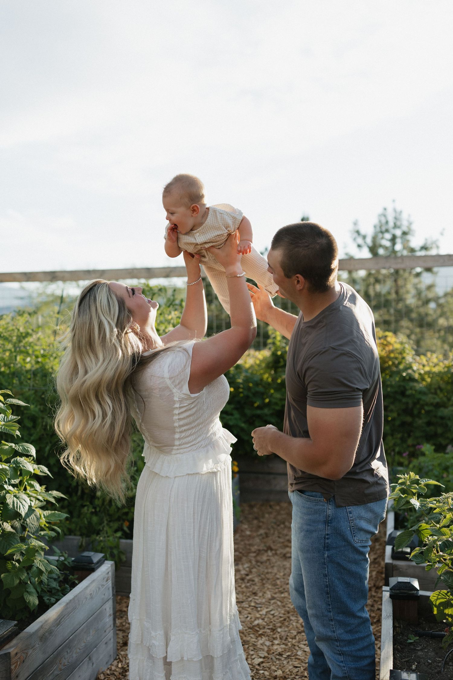 Joyful family moment captured in a garden setting with raised baby against sky.