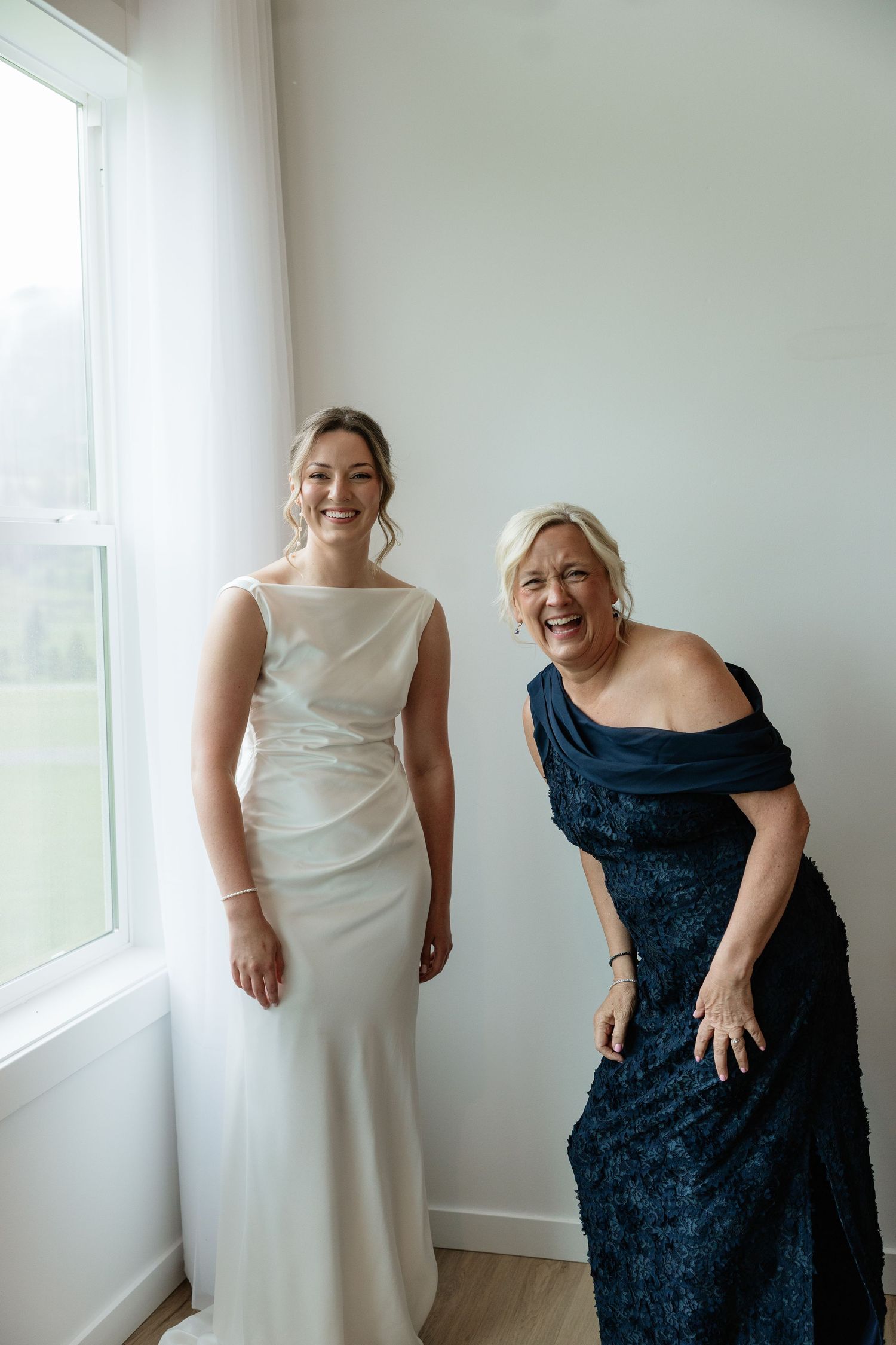 Bride in white gown and woman in navy blue dress laughing together near a window on wedding day.