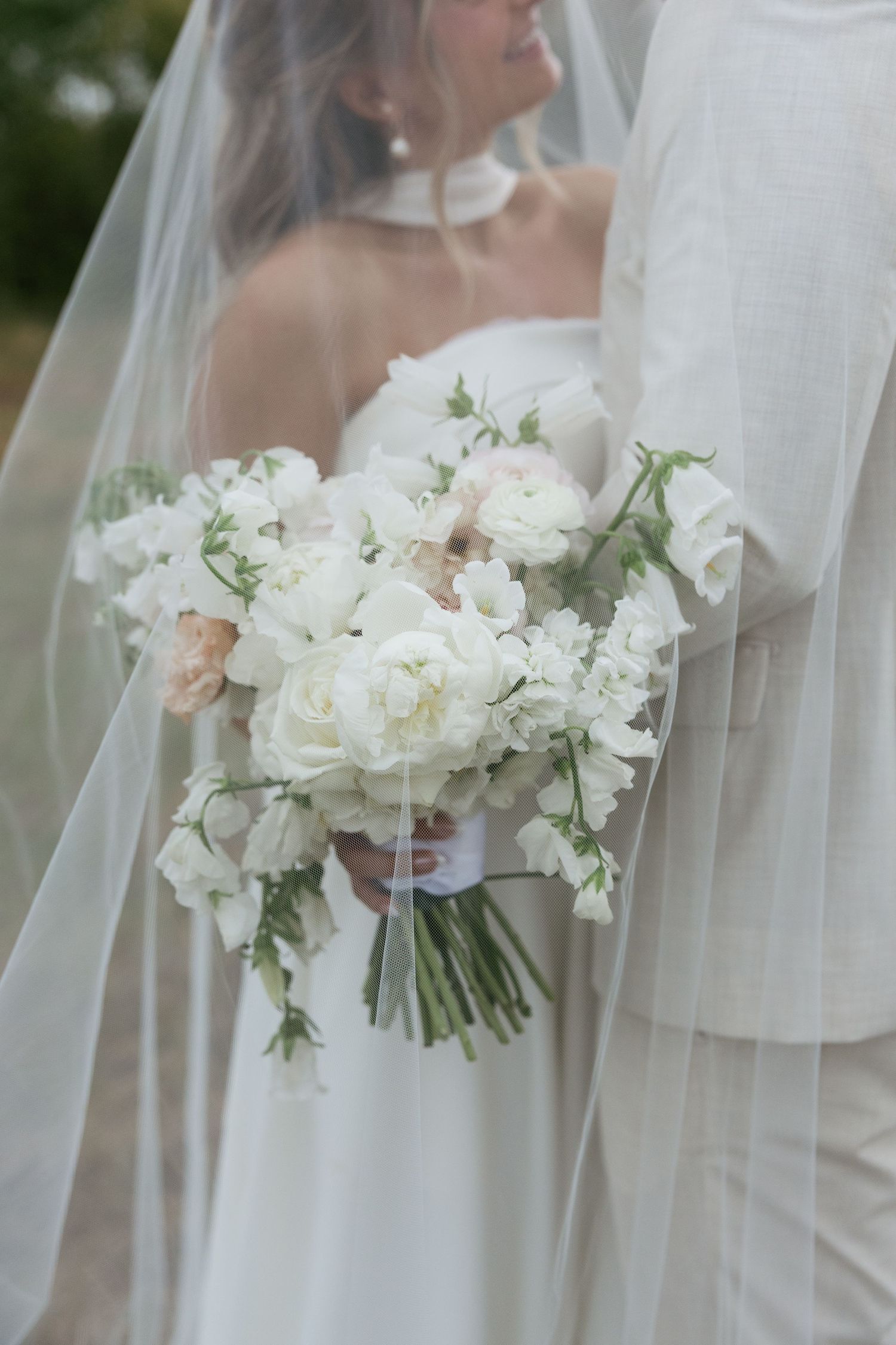 Bride holding a lush white peony and sweet pea wedding bouquet, wearing a flowing veil and strapless gown.