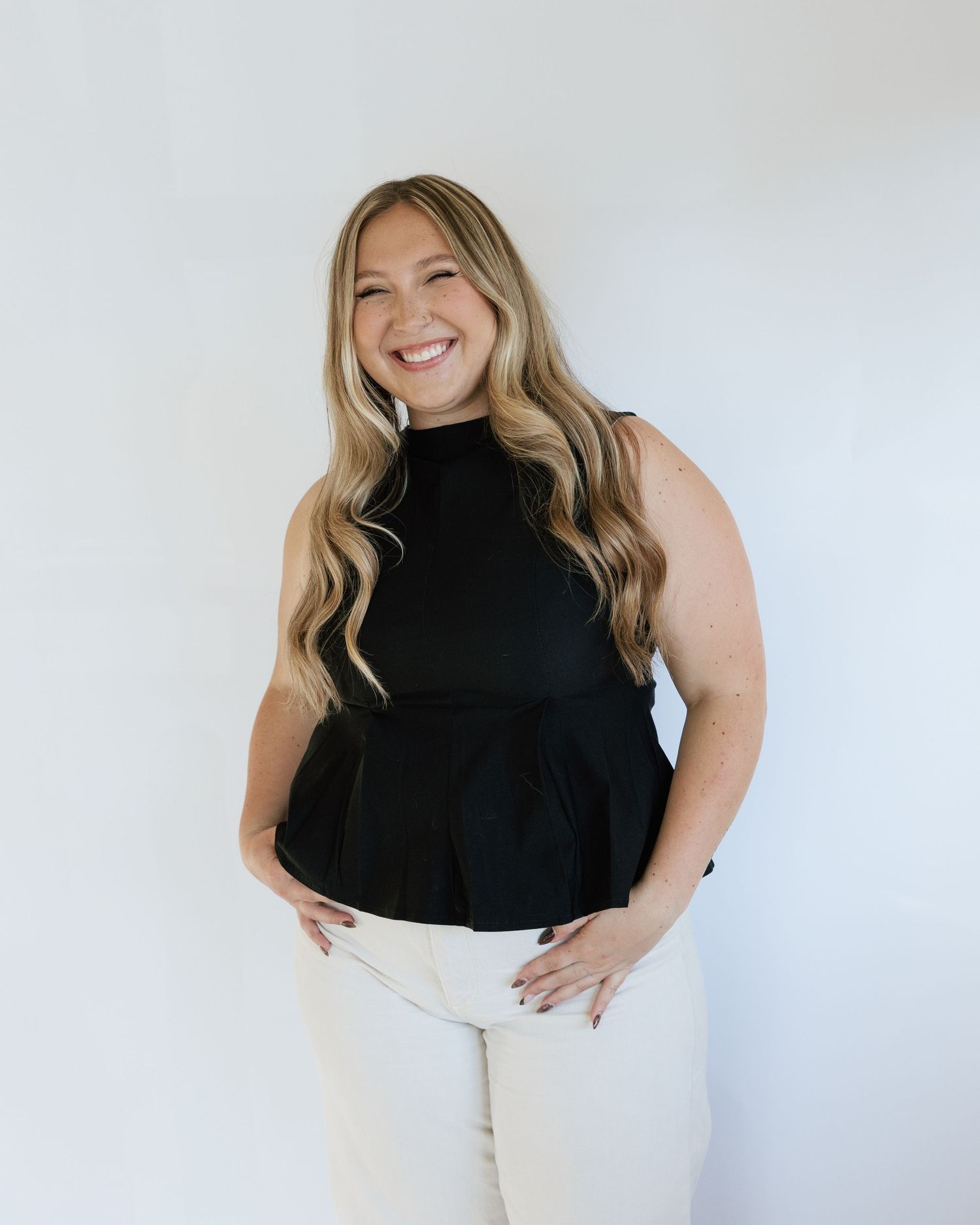 Woman smiling in black sleeveless peplum top and white pants against white background.