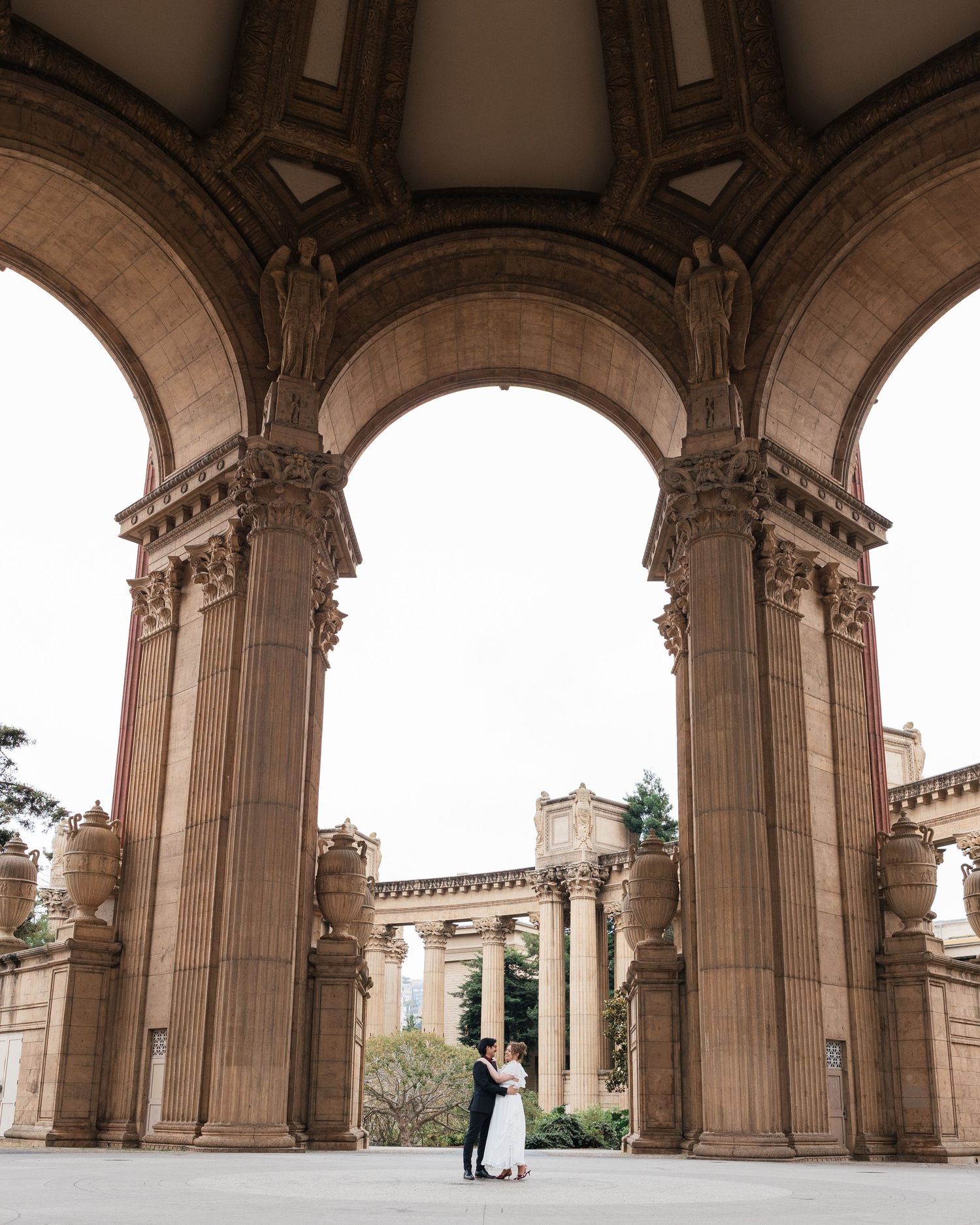 Couple embracing under grand arched colonnade at Palace of Fine Arts, San Francisco, in wedding attire.
