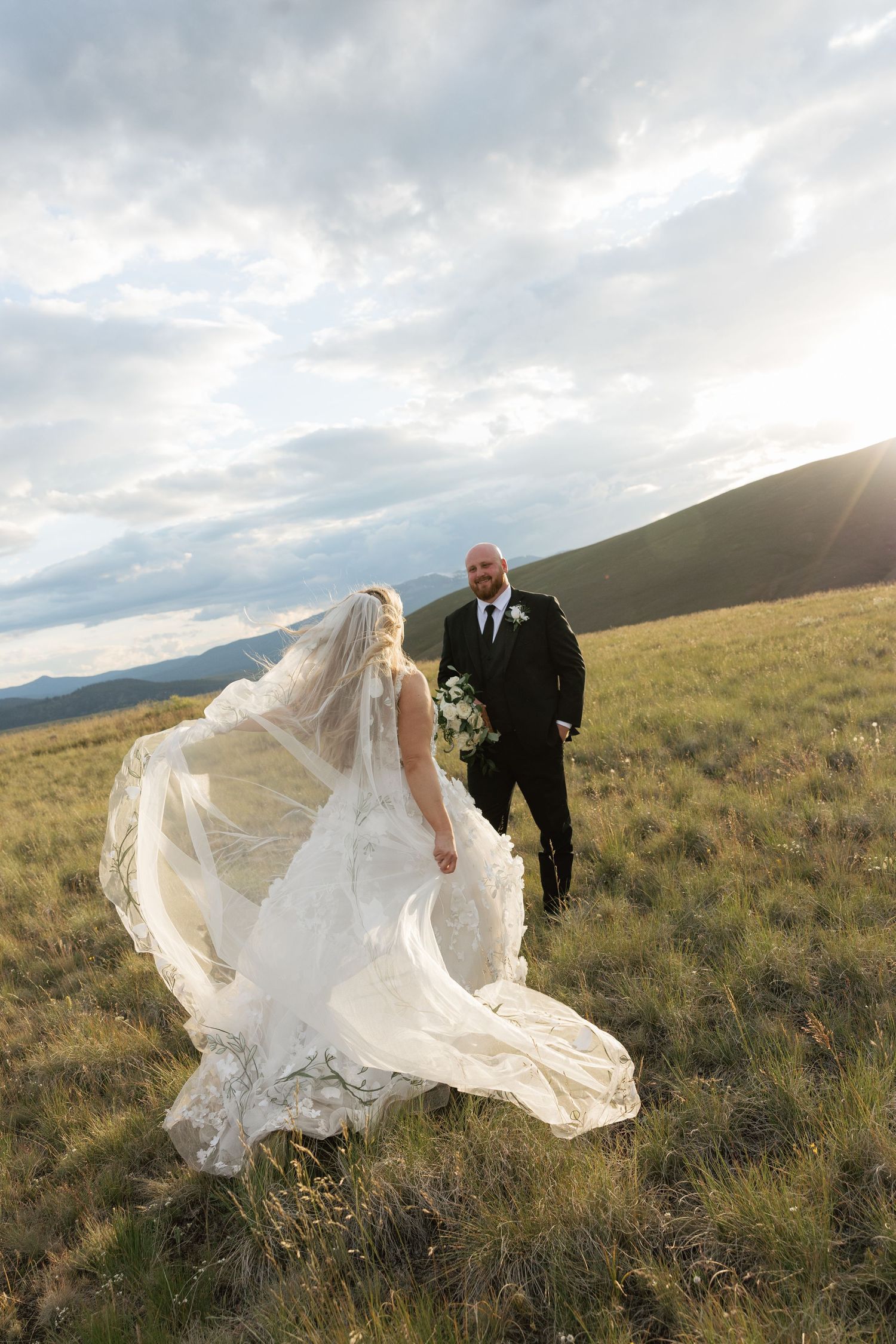 Bride in flowing lace gown and veil walks toward groom in black suit on a scenic mountain meadow at sunset.