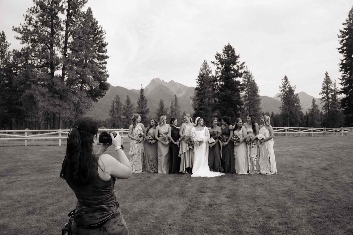 Photographer capturing wedding party with bridesmaids and bride posing outdoors with mountain and pine tree backdrop.