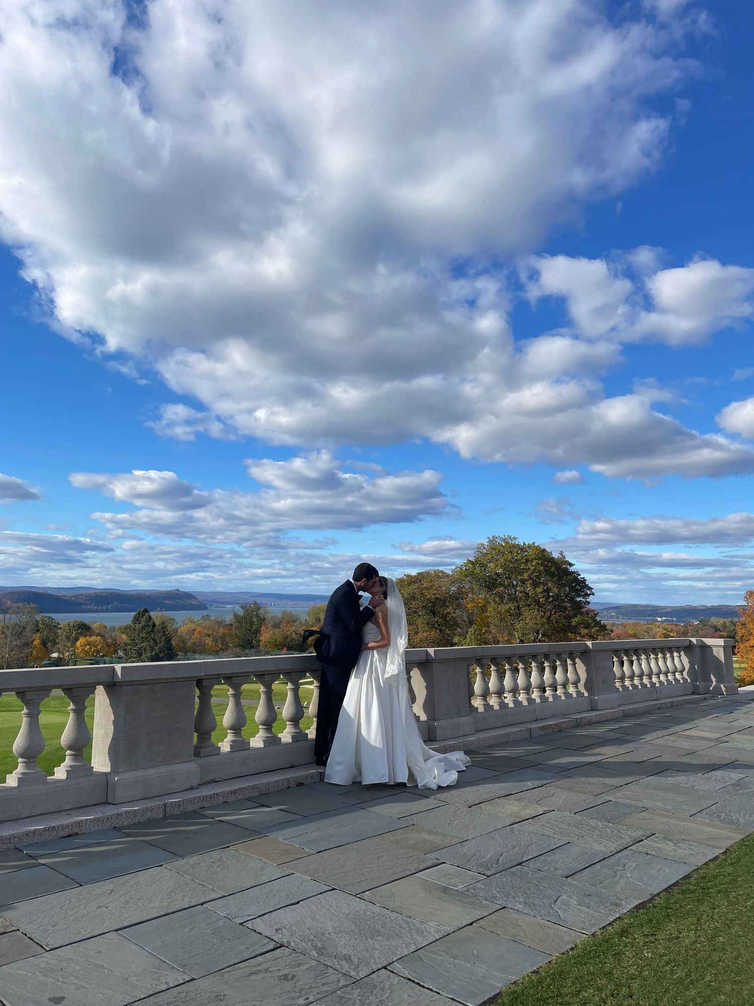 Bride and groom kissing on stone terrace with scenic river valley and dramatic cloudy blue sky backdrop.