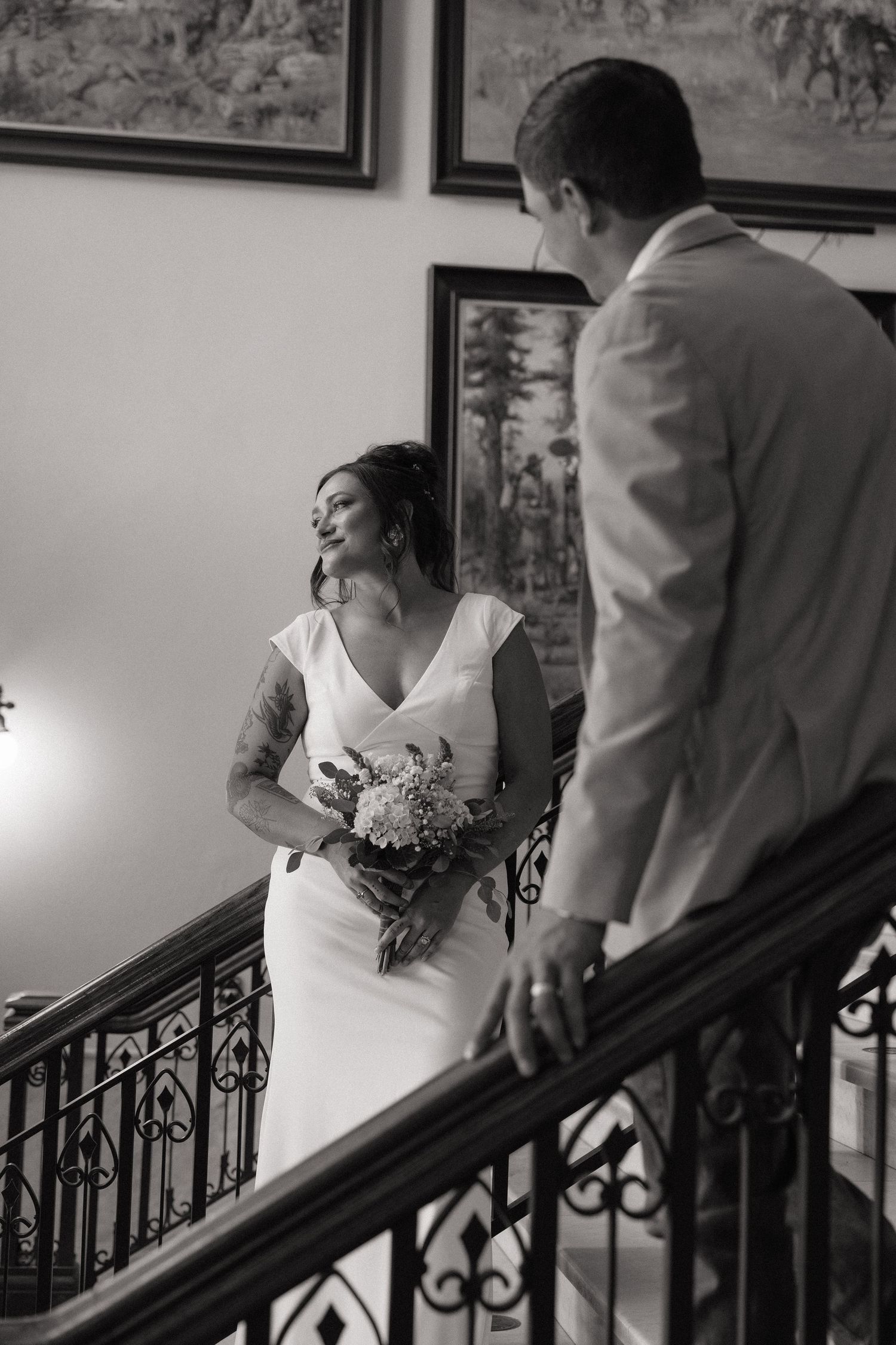 Black and white photo of a bride holding a bouquet on a staircase, smiling at a groom in a white suit.