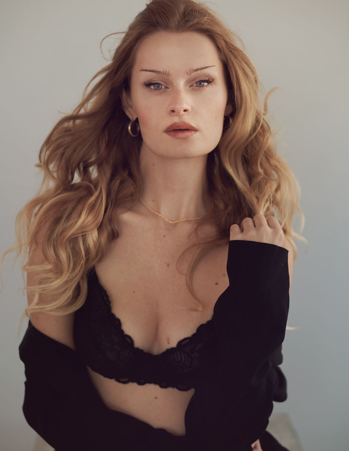 Young woman with long wavy blonde hair wearing black lace bralette and cardigan posing against white background by Jr Francisco Photography