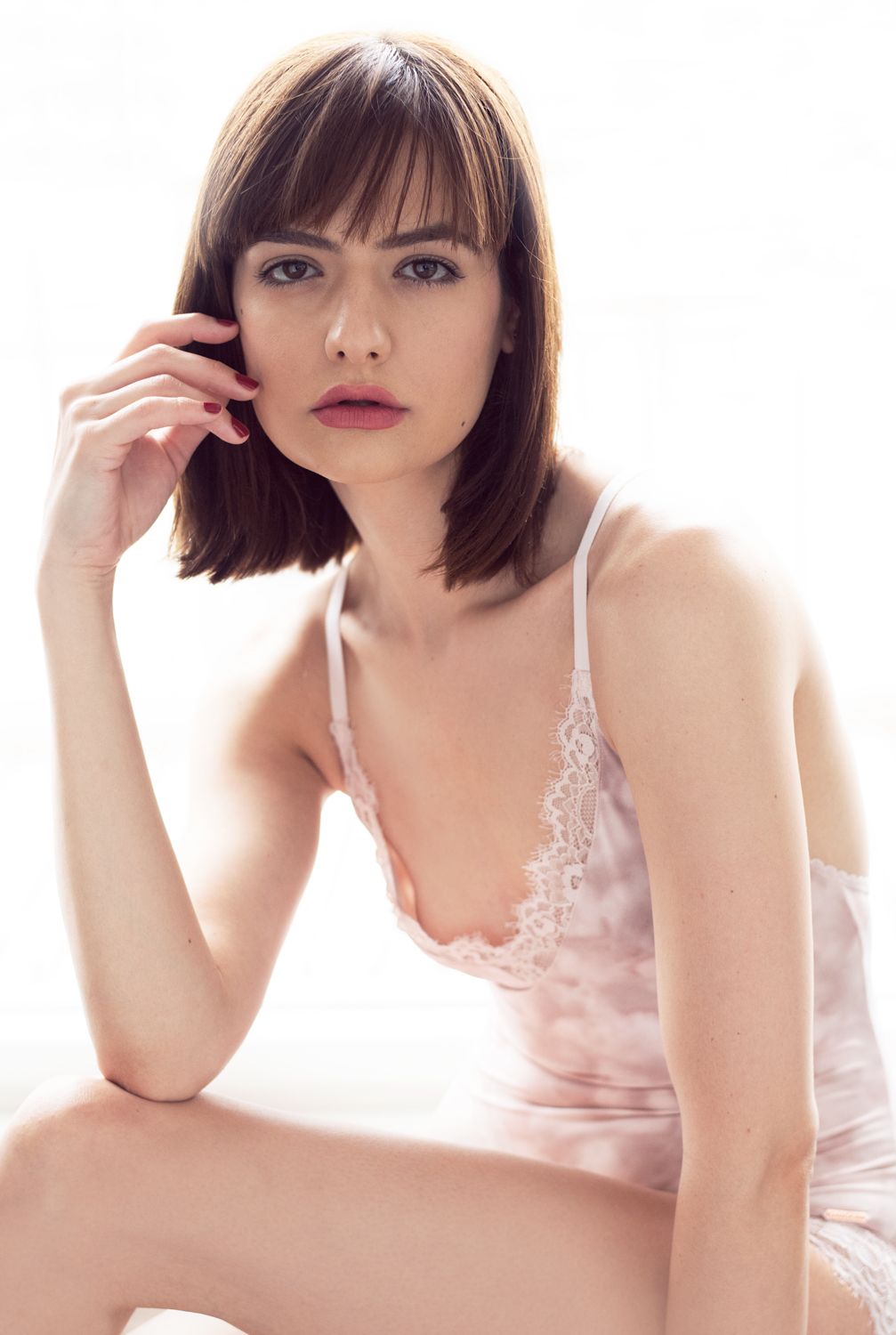 A portrait of a woman by Jr Francisco with dark bob haircut wearing a pink lace slip dress, posing elegantly against a white background.