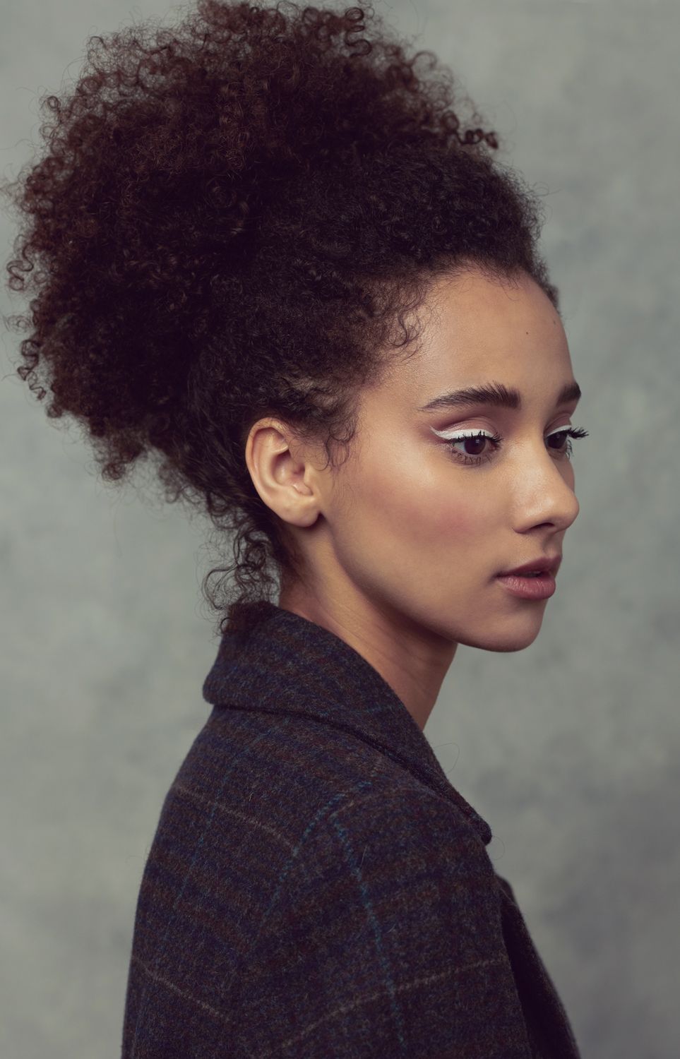 Portrait of a woman by Jr Francisco with natural curly updo hairs wearing a plaid blazer, photographed in profile against a gray background.
