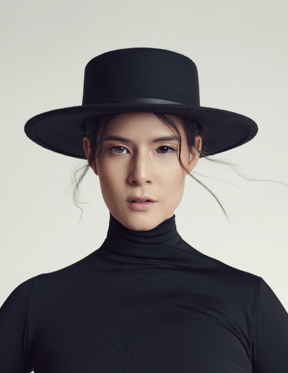 A photo of a woman taken by Jr Francisco in black turtleneck and black hat poses against light background in elegant fashion portrait.
