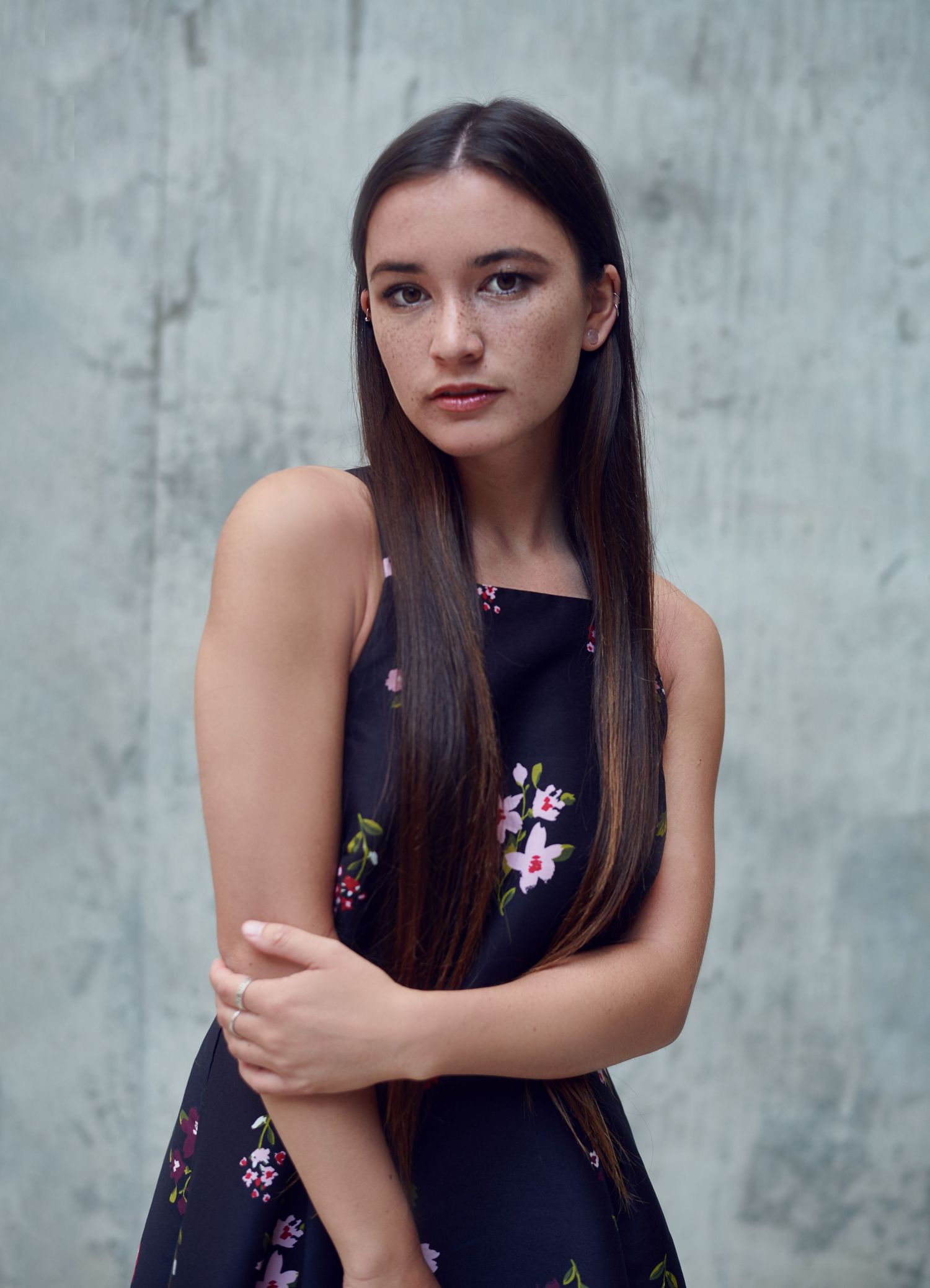 a photo Young  Woman taken by Jr Francisco with long dark hair and freckles wearing a black floral dress, posing against a gray wall.