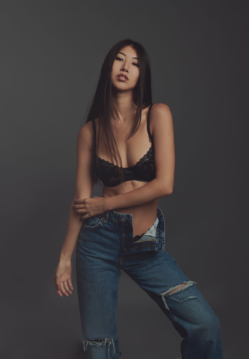 photo of a woman taken by Jr Francisco in black lace bralette and distressed blue jeans posing confidently against dark gray background.