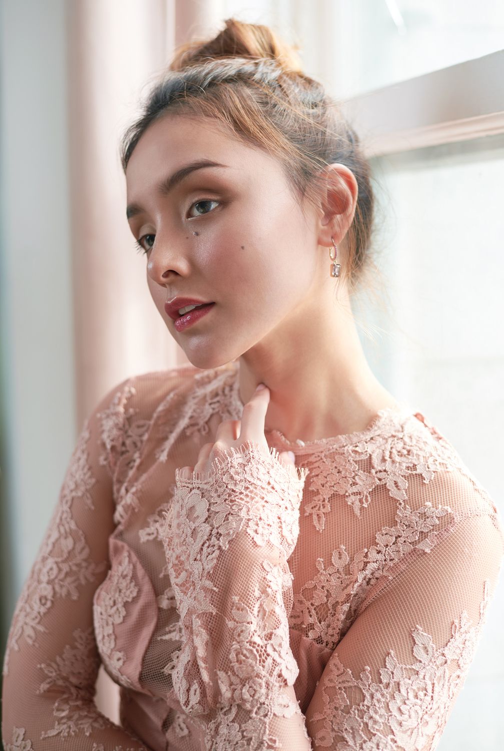 Portrait of a Young woman by Jr Francisco in pink lace long-sleeve dress with braided updo hairstyle and drop earrings posing near a window.