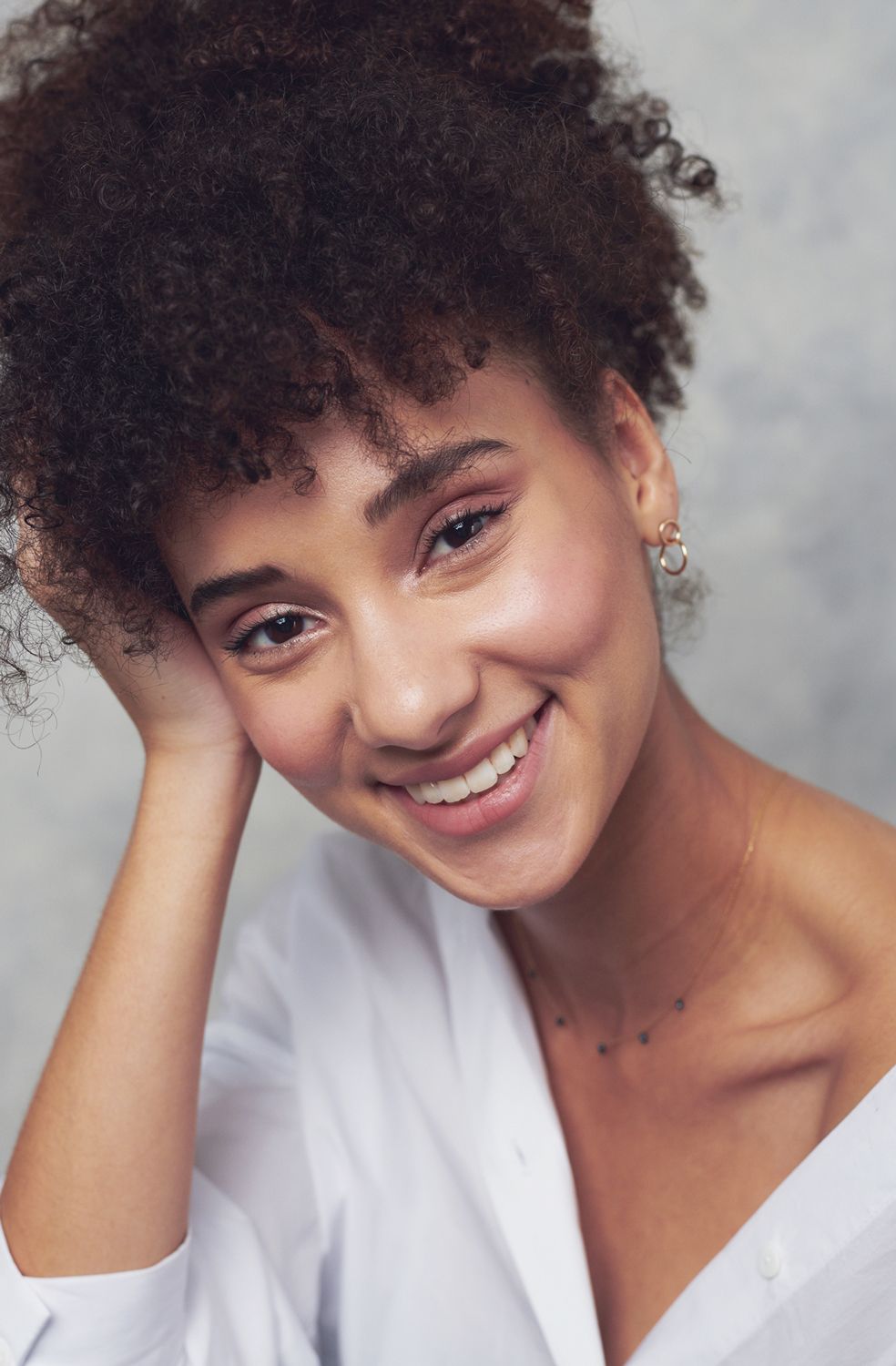 photo of a Smiling young woman by Jr Francisco with curly hair wearing a white shirt, resting her hand on her head against a gray background