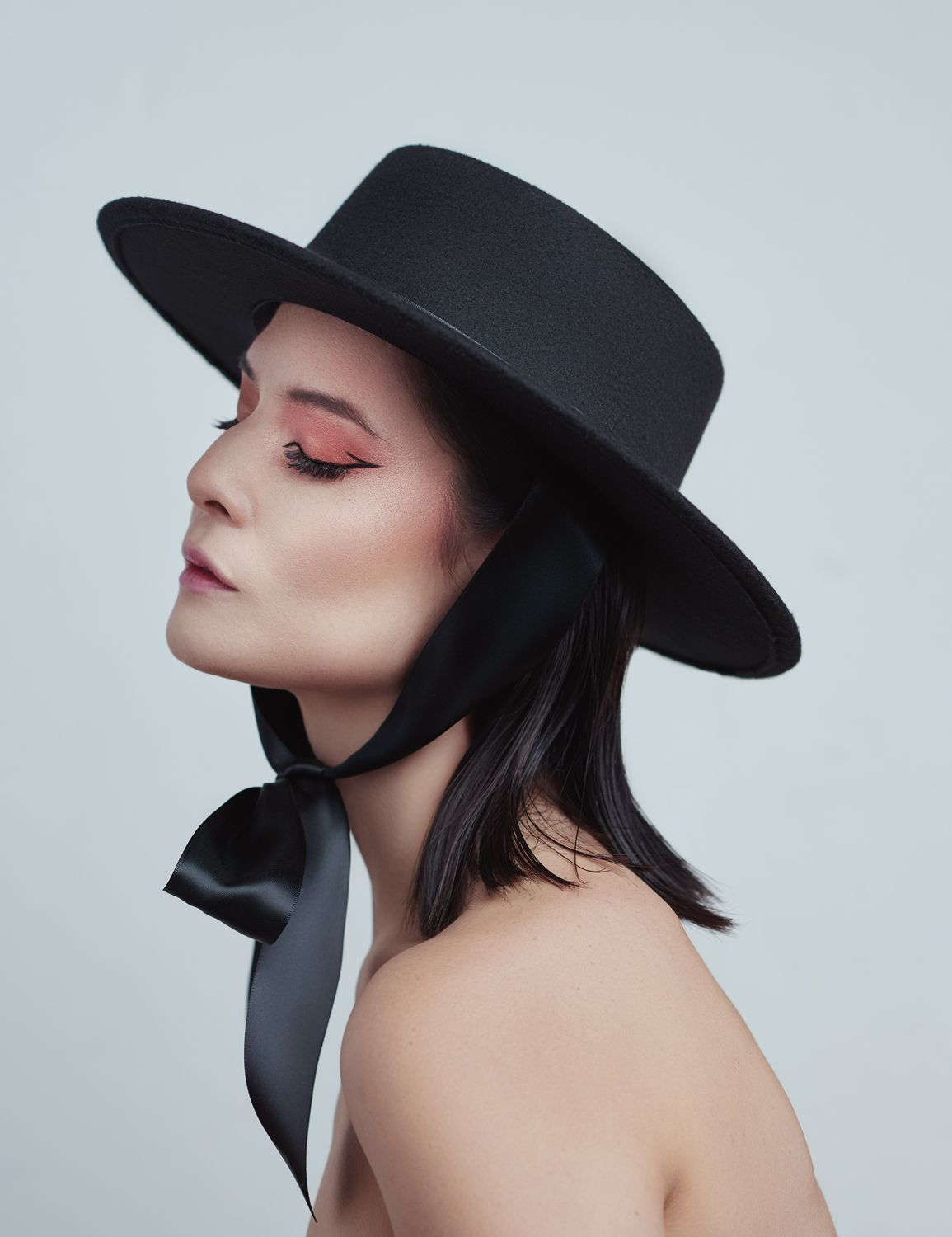 Woman in profile wearing a black wide-brim hat with satin ribbon, bold pink eye makeup, against a light background.  Photo by Jr Francisco.