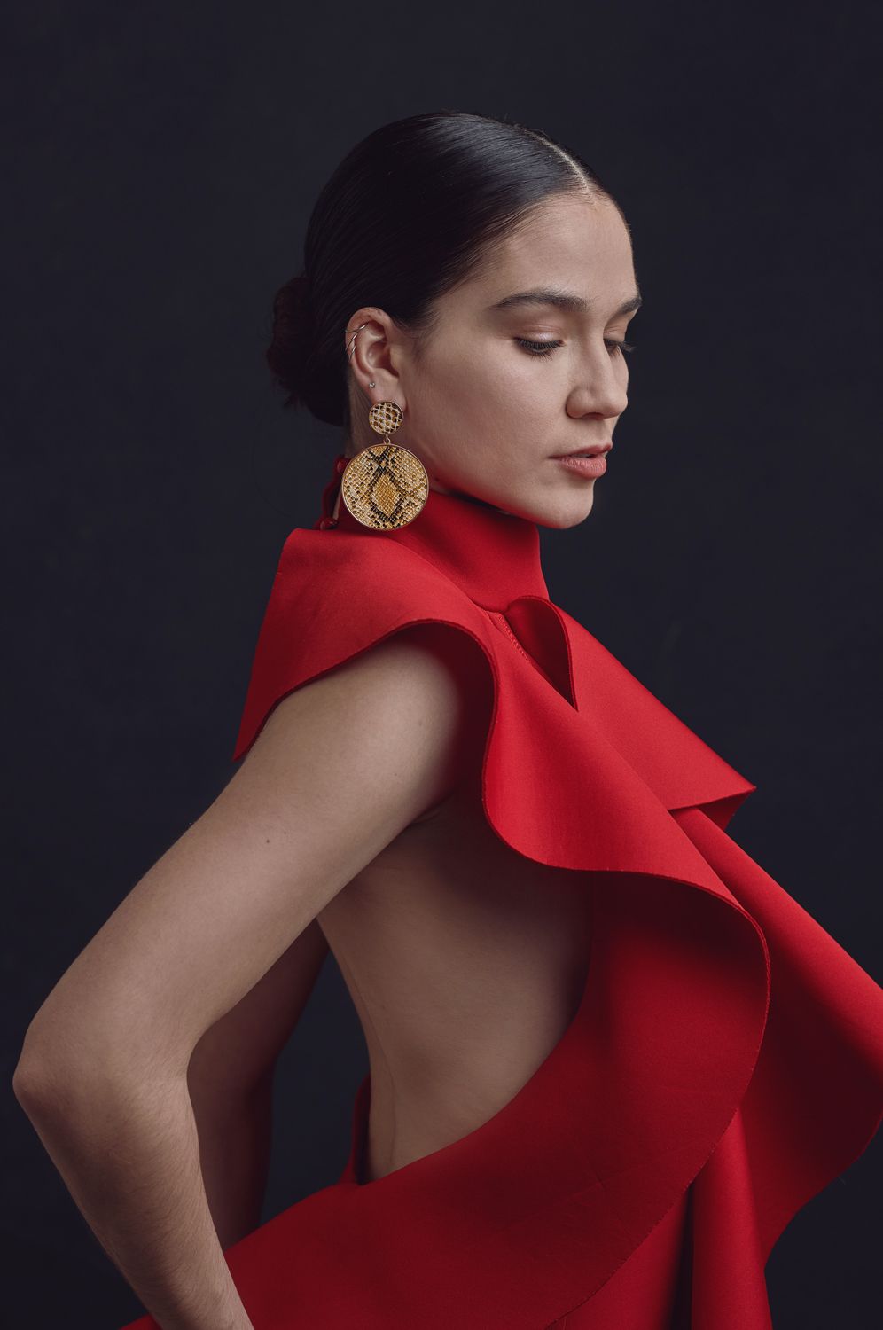Photo of a Woman taken by Jr Francisco Photography in red backless dress with gold earrings poses elegantly against dark background.