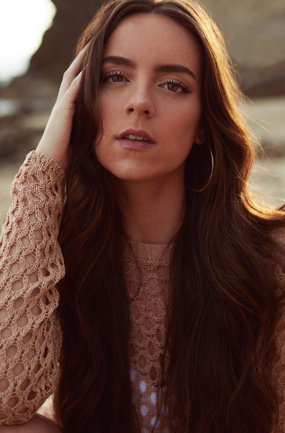 A portrait of a woman by Jr Francisco with long brown hair wearing a lace top and hoop earrings poses outdoors near rocky cliffs.