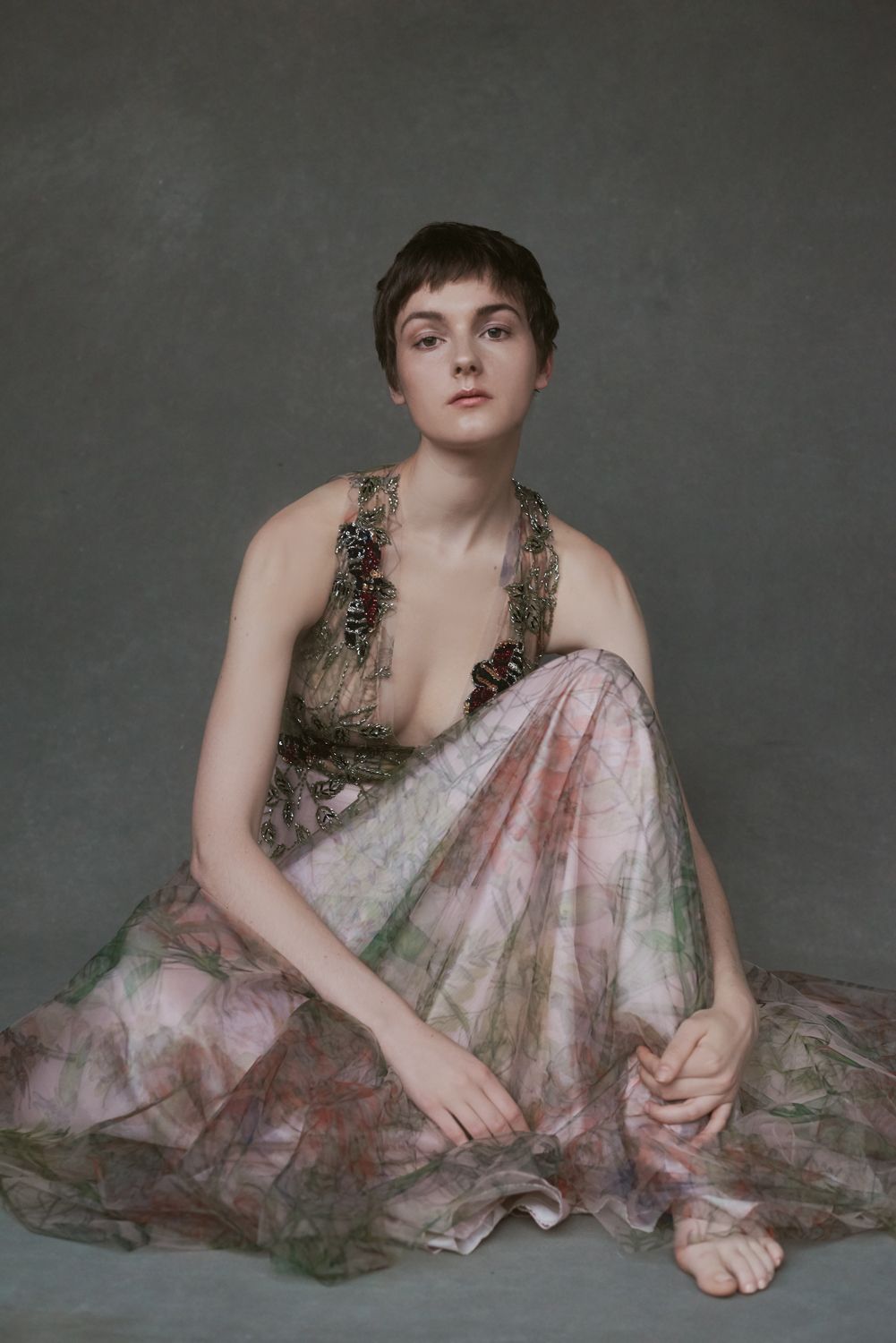 Portrait of a woman by Jr Francisco with short hair sits gracefully in a flowing floral gown with jeweled neckline against gray backdrop.
