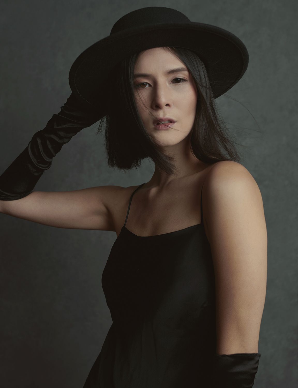 a photo by Jr Francisco of a Woman in black dress, wide-brim hat, and long gloves poses elegantly against a dark studio background.