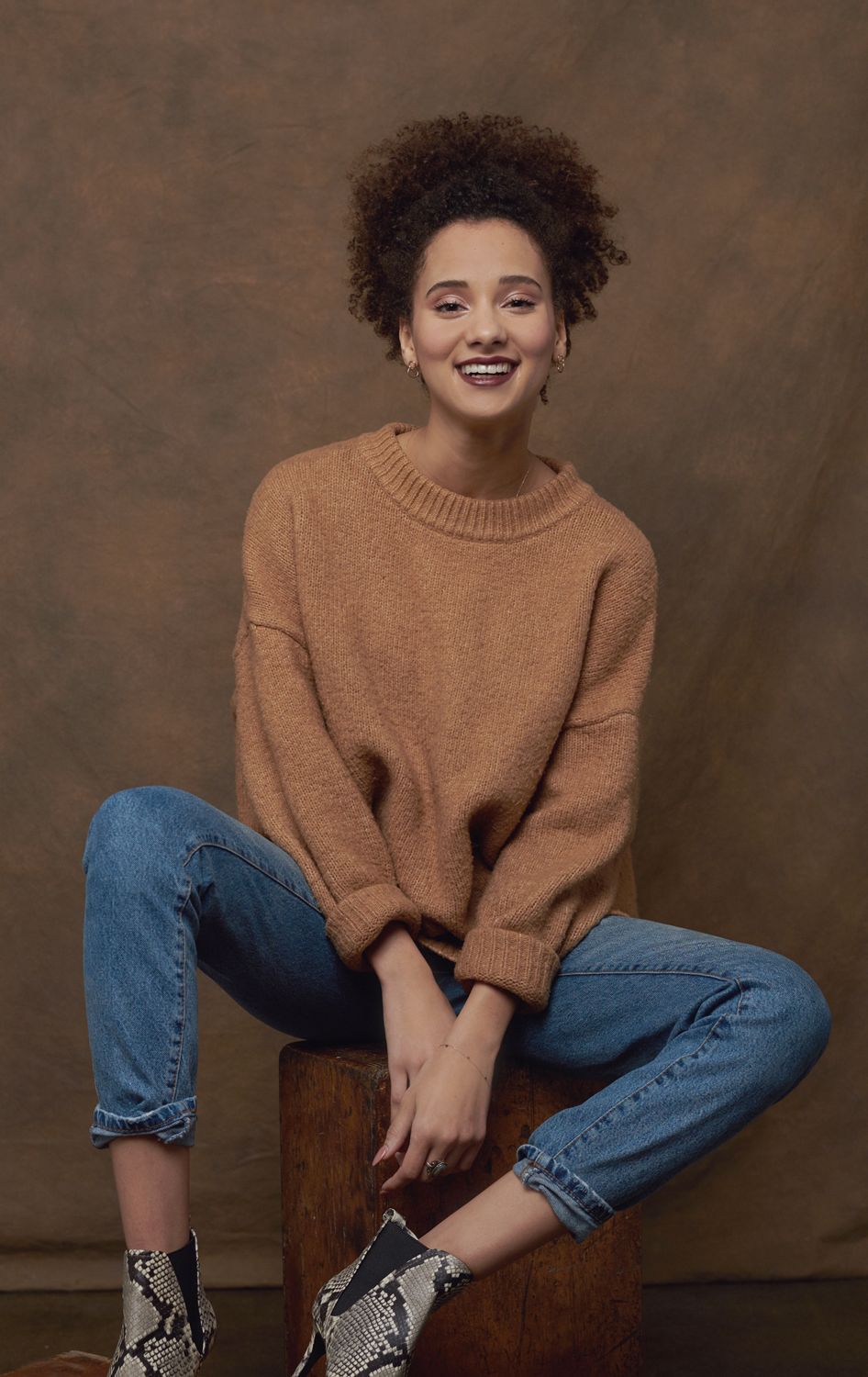 Photo of a Young woman by Jr Francisco in camel knit sweater and blue jeans sits smiling, wearing snakeskin boots against brown backdrop.