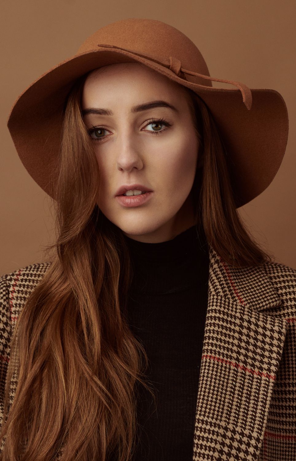 Photo of a Young woman by Jr Francisco in brown floppy felt hat and plaid blazer posing against a warm beige background.